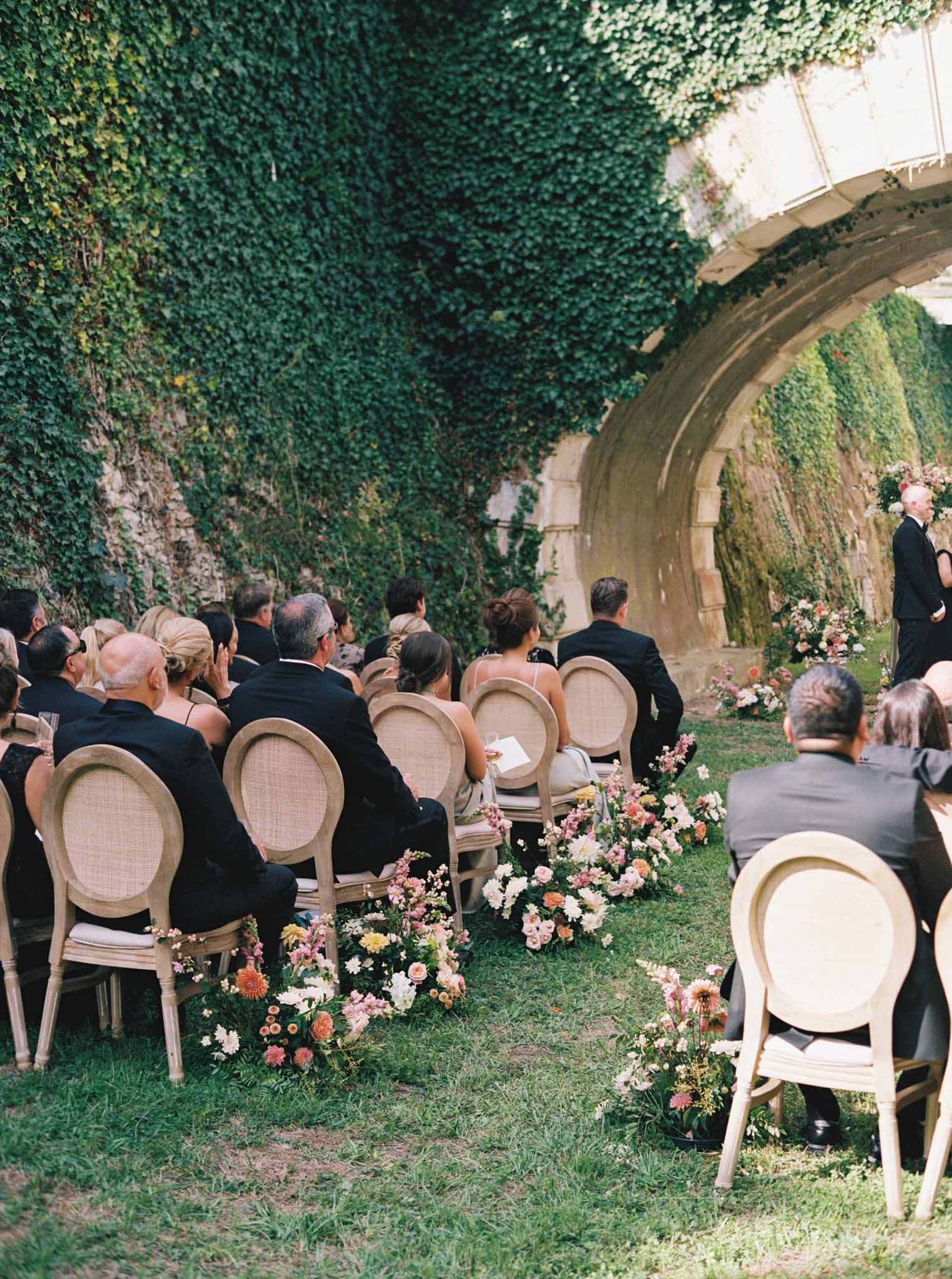 Outdoor wedding ceremony in ivy-covered stone courtyard with guests seated facing archway and coral pink aisle flowers