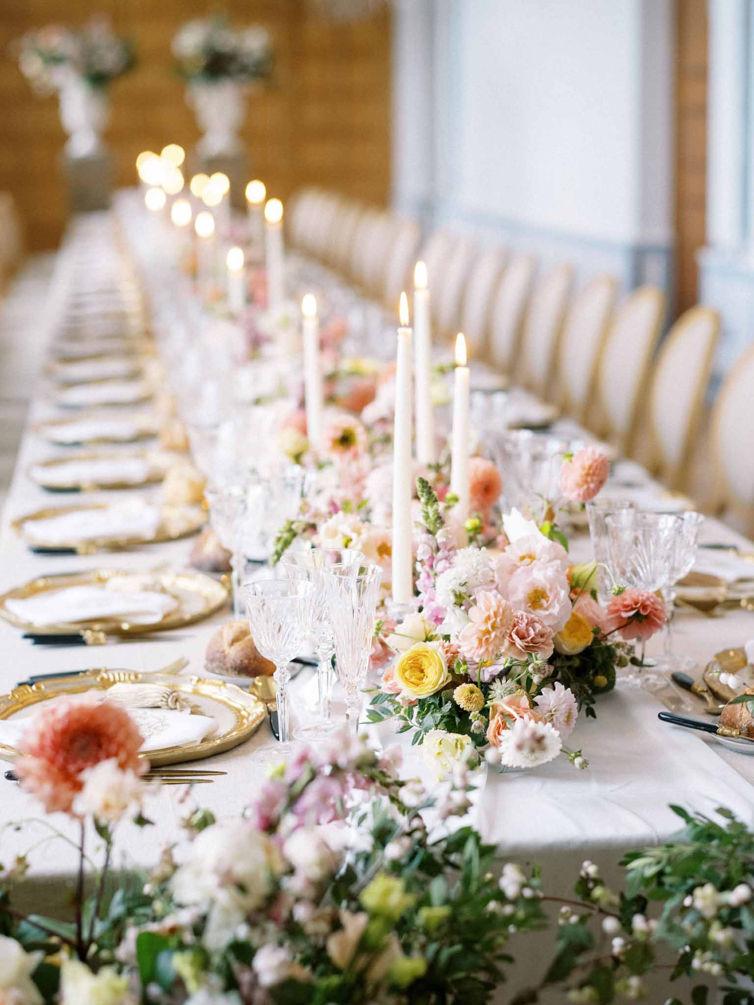 Long reception table with coral roses, yellow flowers, and gold-rimmed chargers, taper candles in candleholders