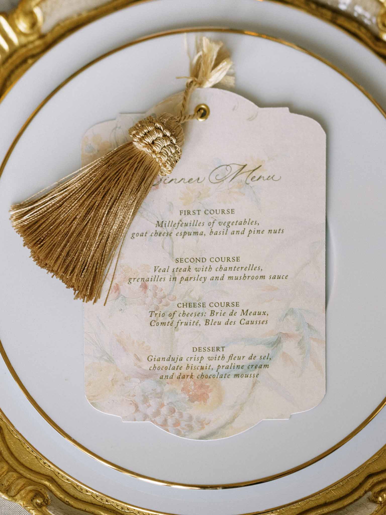 Watercolor floral menu card with gold tassel on gold-rimmed charger plate at formal place setting