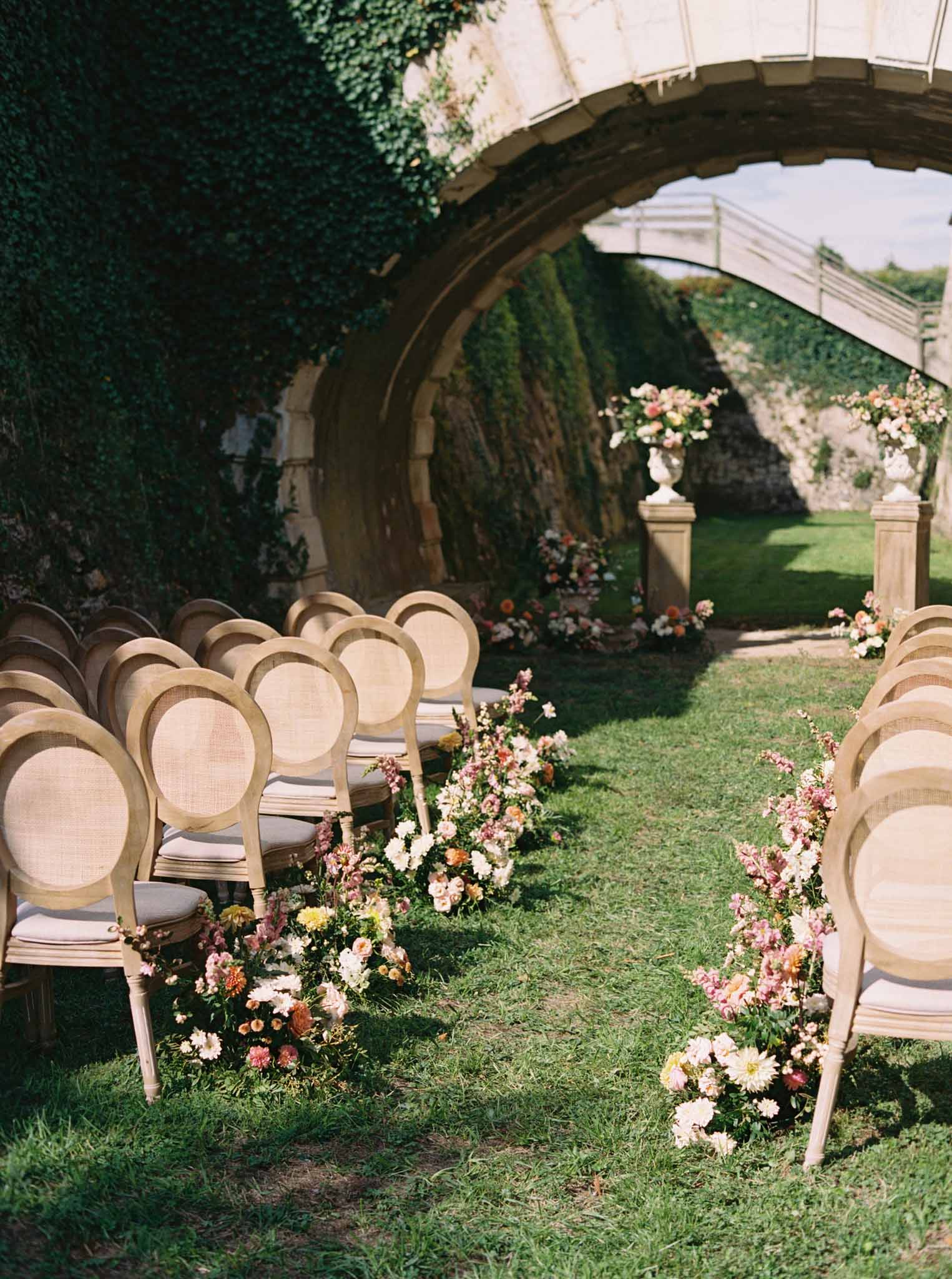 Outdoor ceremony in walled garden with stone archway, rattan chairs, blush and cream aisle floral arrangements