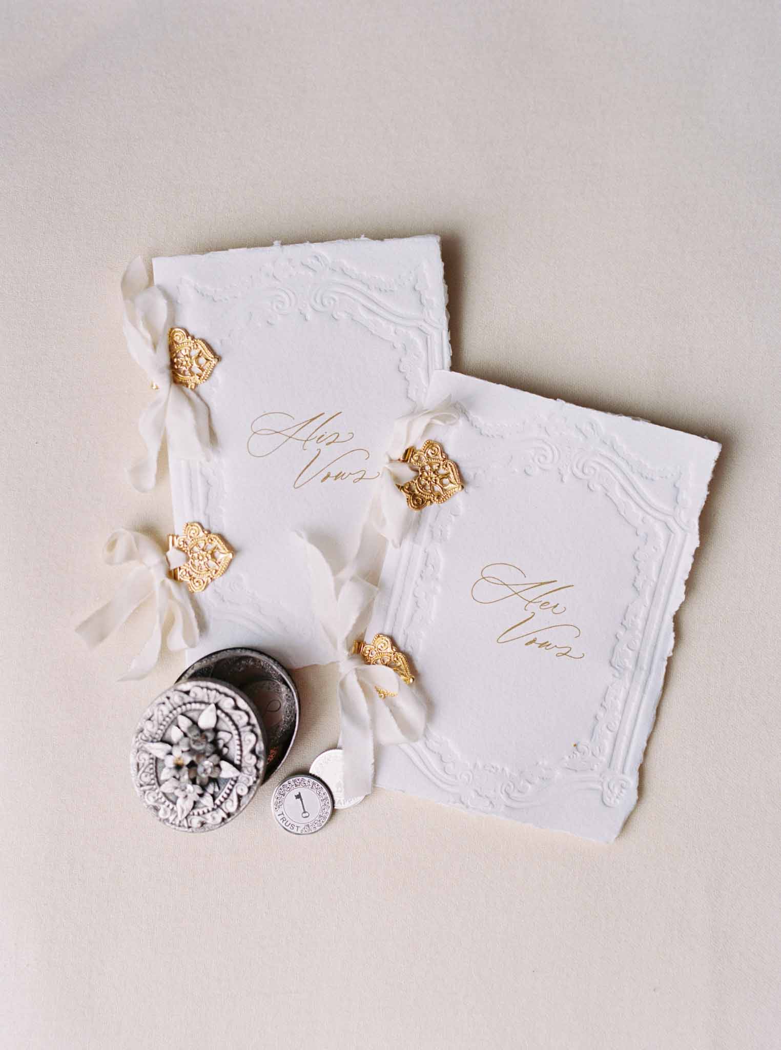Flat lay of wedding stationery with white embossed invitation cards, gold filigree charms, cream ribbon, and wax seal