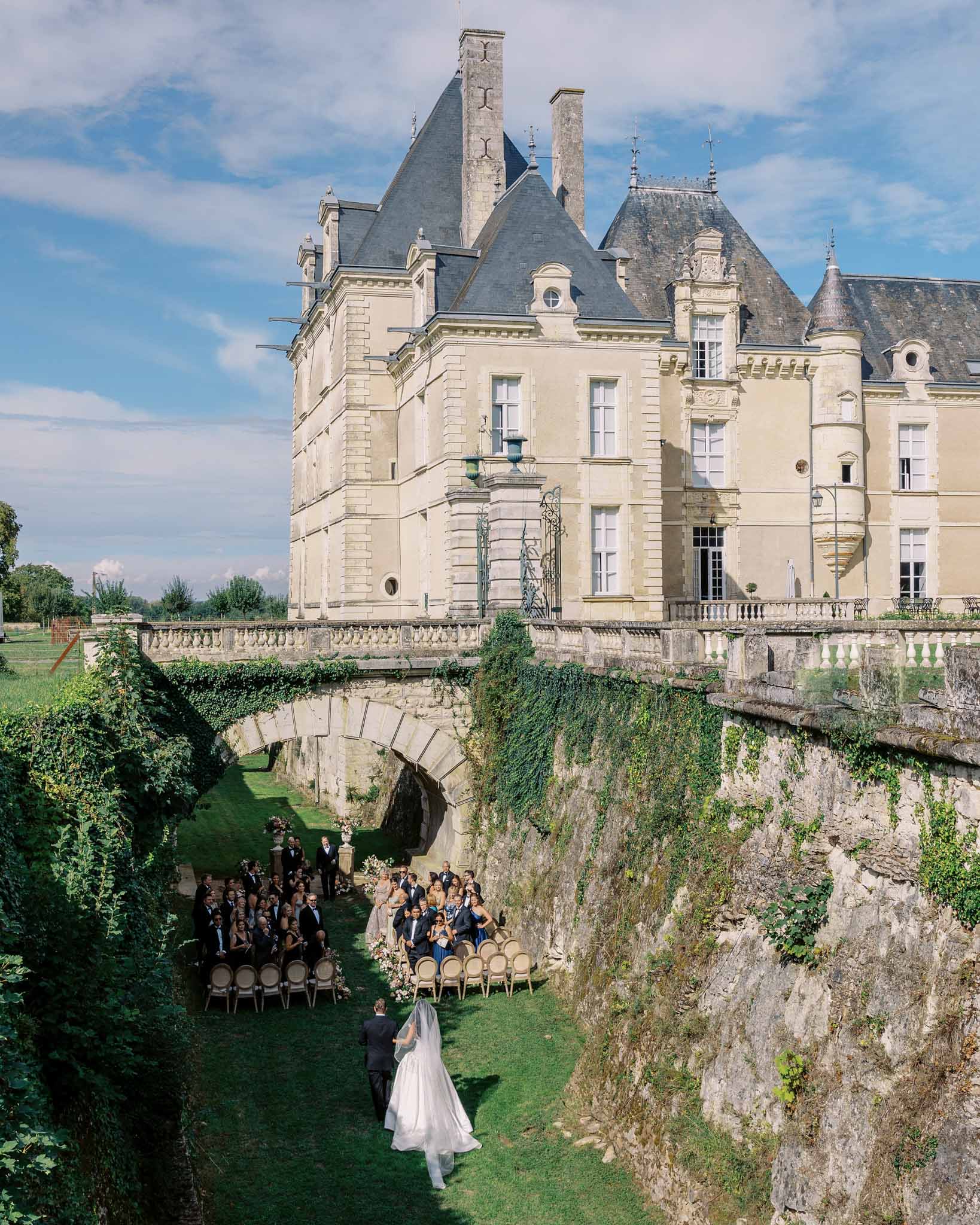 Bride walking toward ceremony in a sunken chateau courtyard with guests seated on wooden chairs below a stone bridge