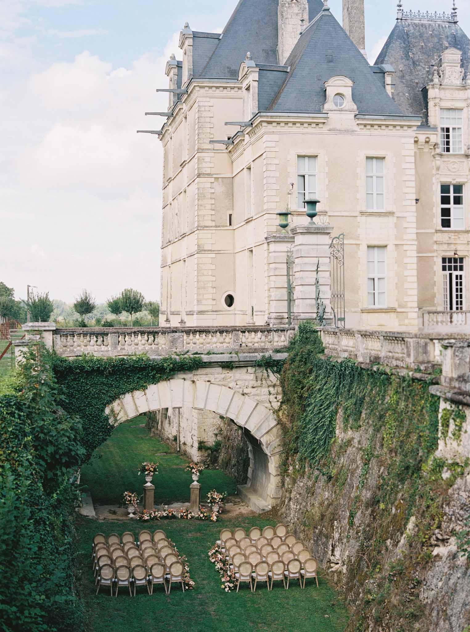 Ceremony setup in chateau courtyard with Chiavari chairs facing stone archway bridge and floral urns, cream facade behind