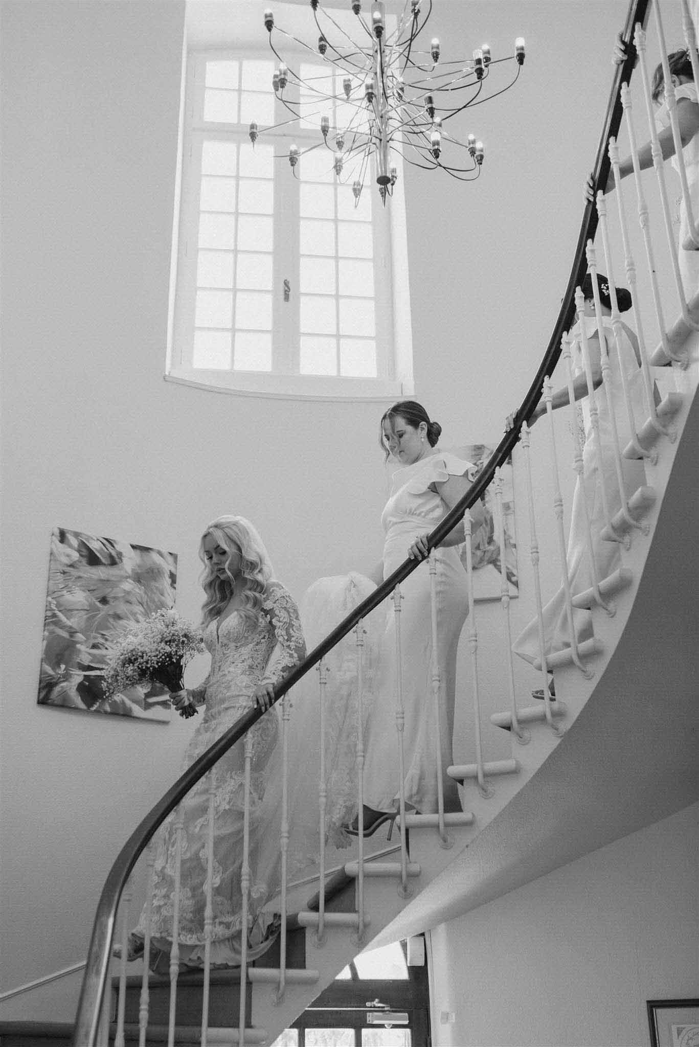 Black and white portrait of bride descending spiral staircase with arched windows and chandelier above