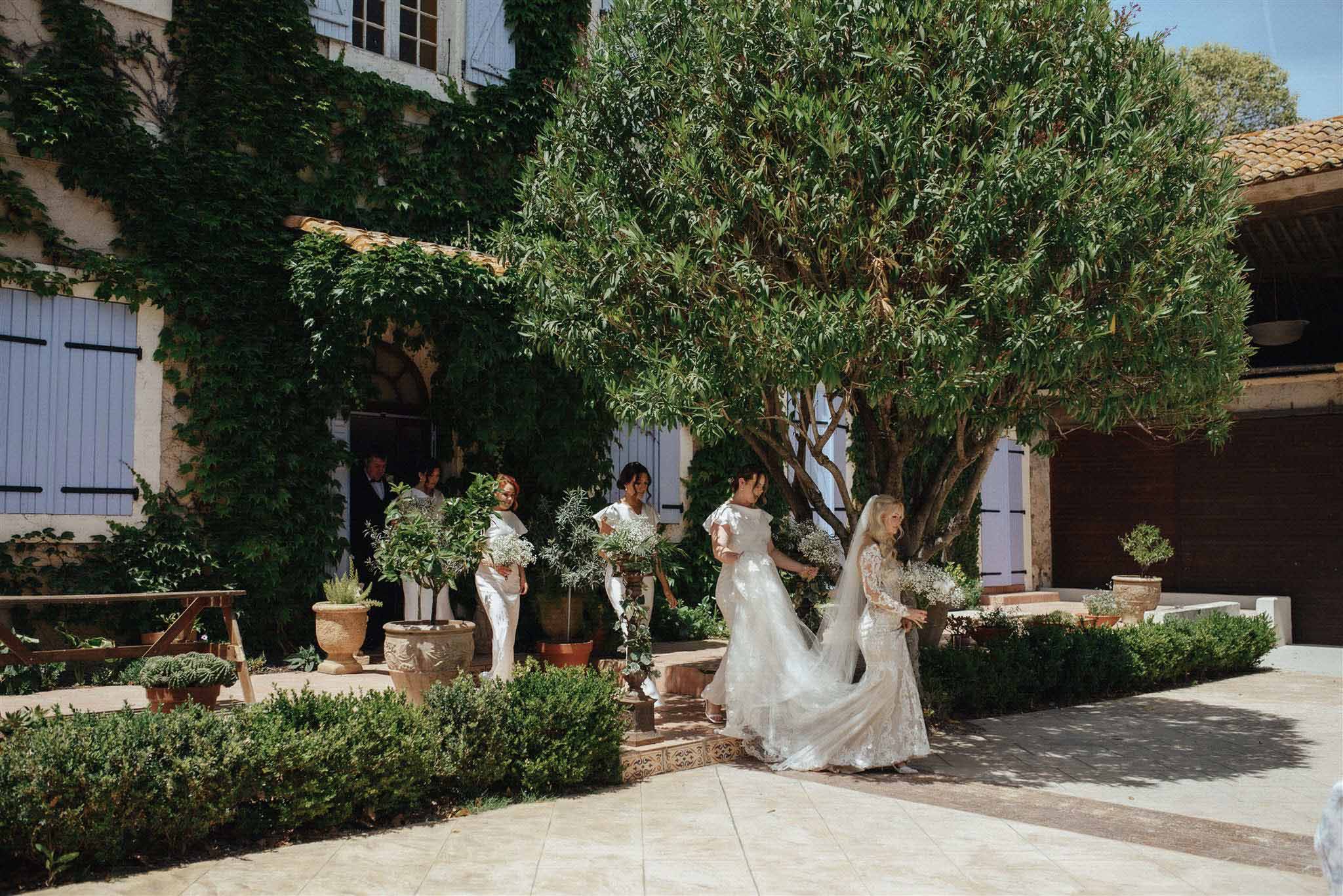 Bride and bridesmaids in ivory lace gowns processing through a Mediterranean courtyard with olive trees and ivy-covered stone walls.