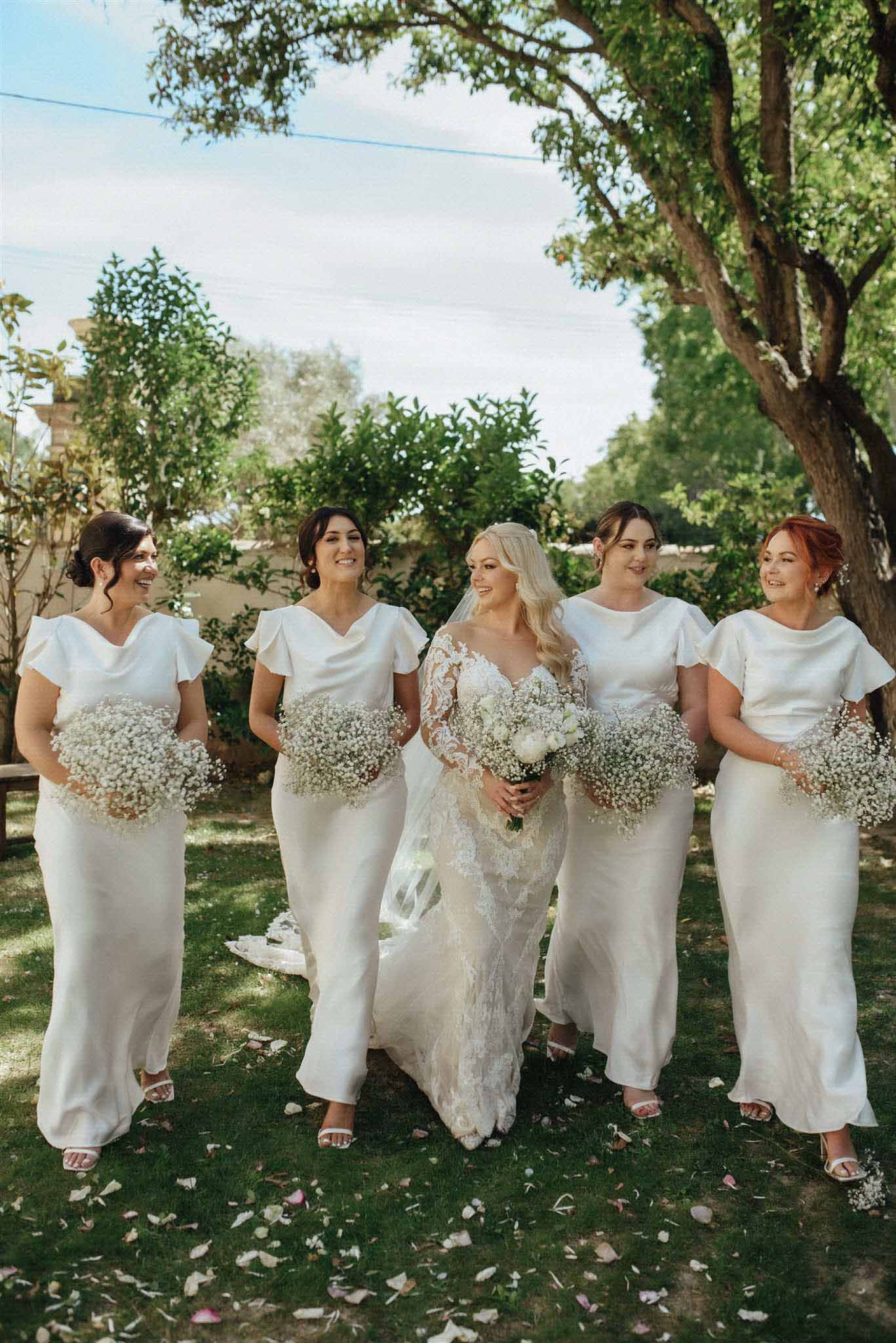 Bride with four bridesmaids in matching ivory slip dresses holding baby's breath bouquets on garden lawn