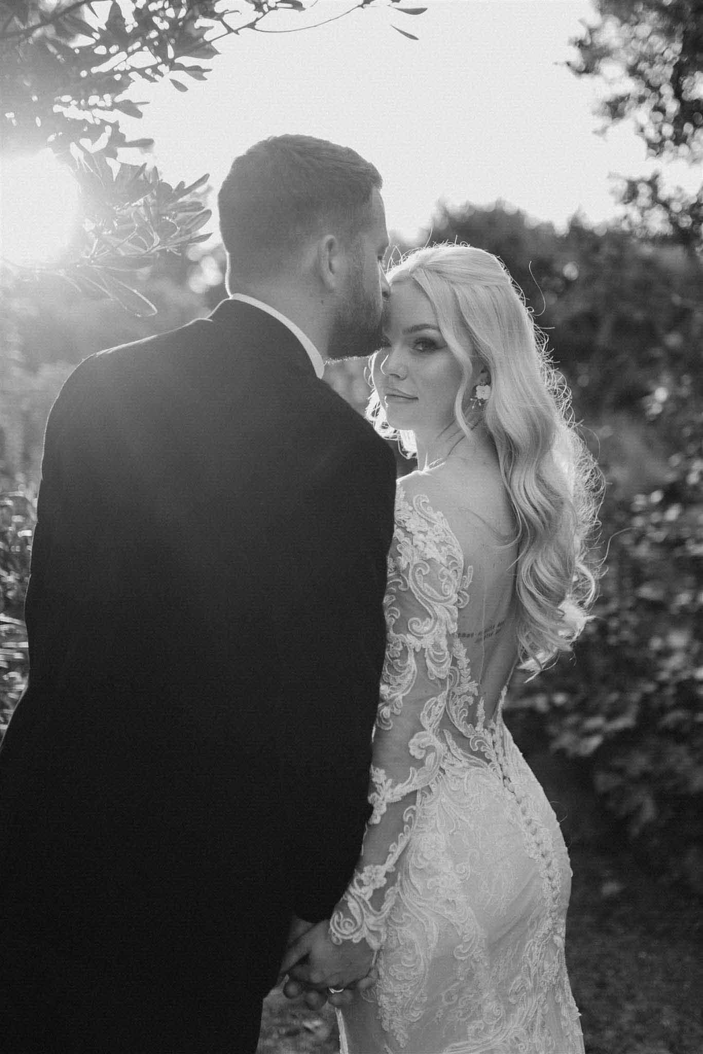 Bride and groom holding hands outdoors, black and white, bride in intricate lace dress with floral vines framing composition