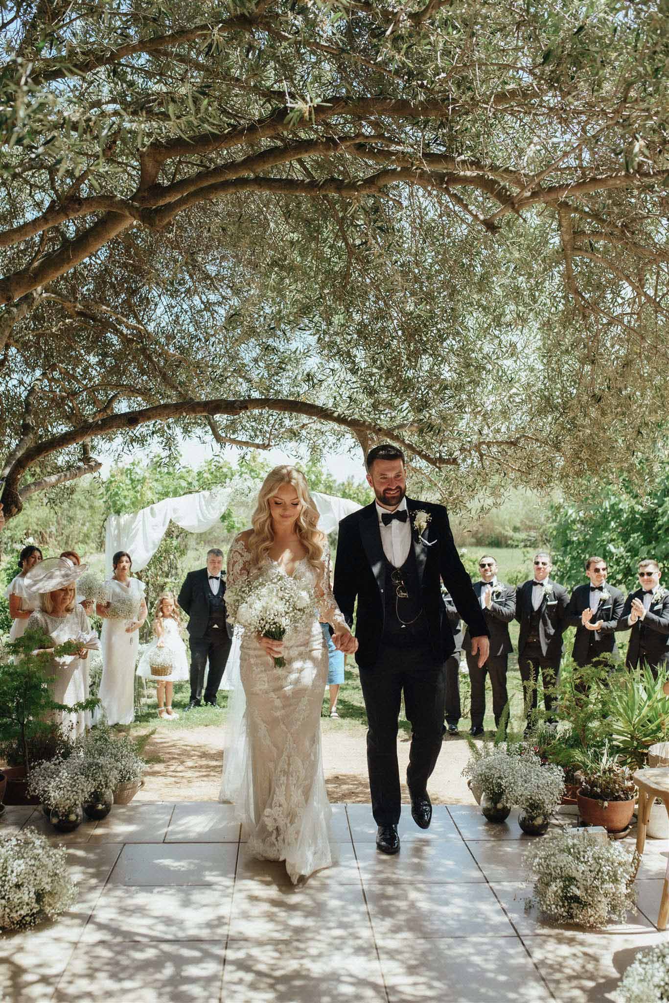 Bride and groom walking the recessional beneath a gnarled olive tree in a Mediterranean courtyard, guests standing on each side.