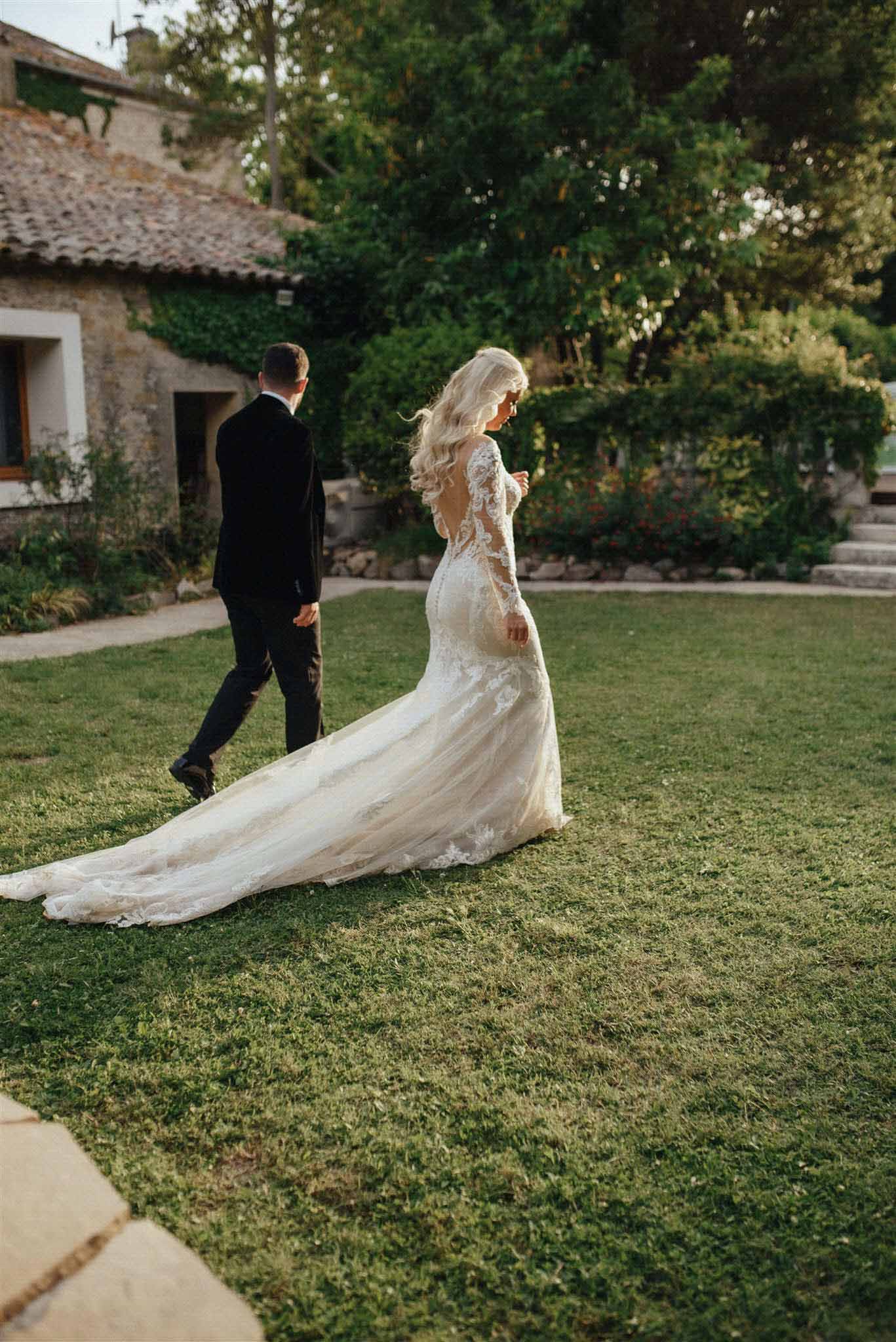 Bride and groom walking across lawn with cathedral train trailing at stone courtyard venue