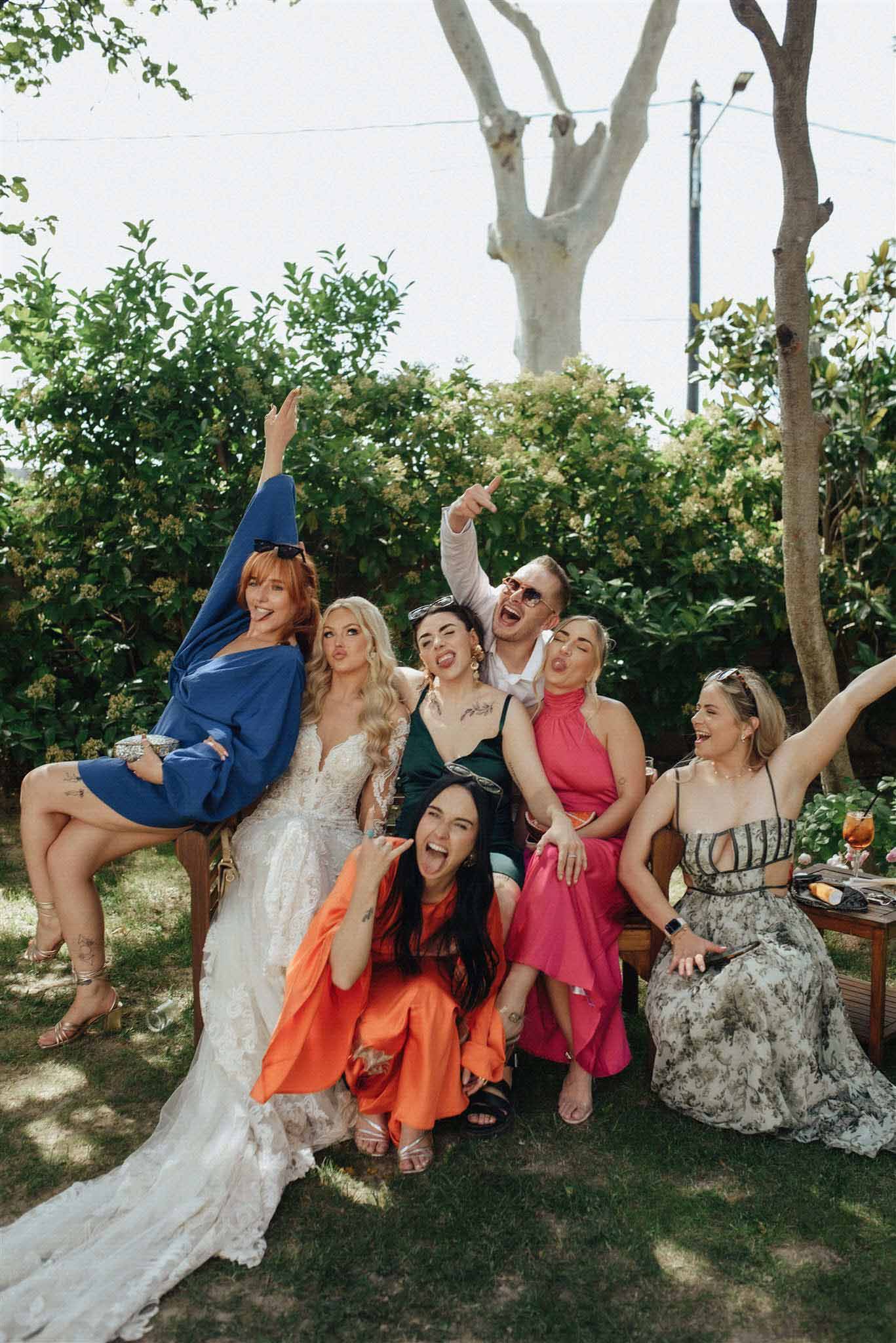 Bride in cream lace gown posing with guests in colourful mix of outfits under garden trees with drinks