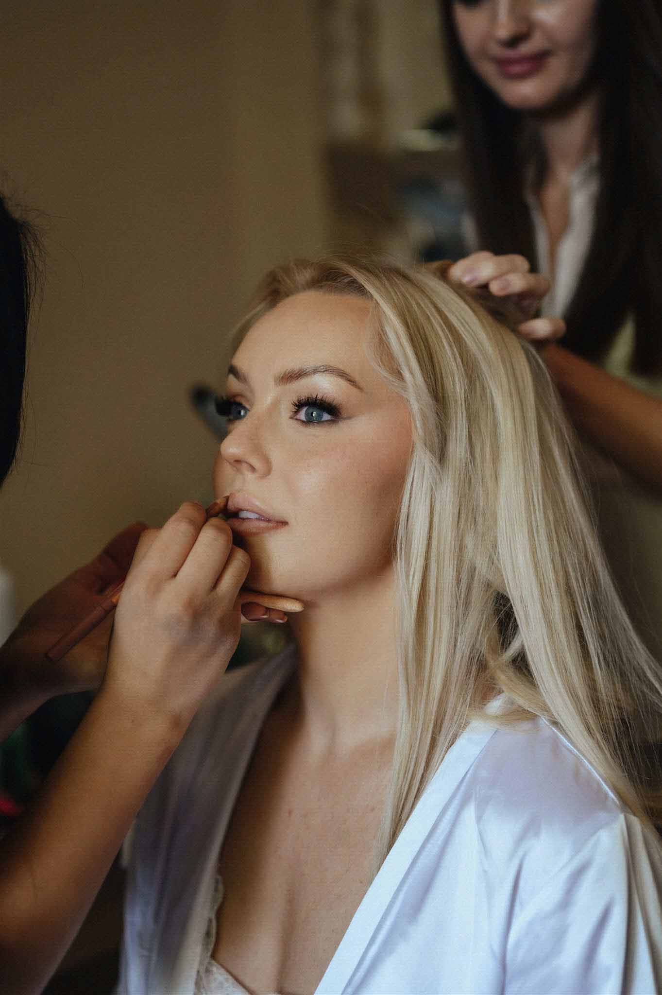 Makeup artist applying lipstick to bride in a white robe with platinum blonde hair in an indoor bridal preparation setting.