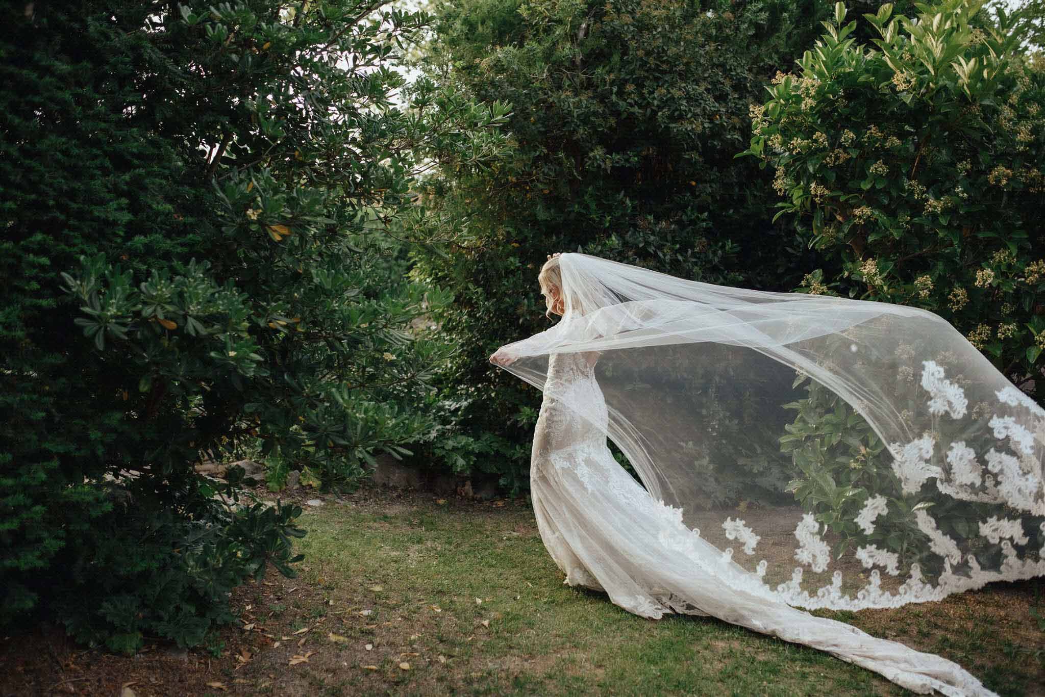 Bride in ivory lace gown with cathedral-length veil posing in a manicured garden with green hedging