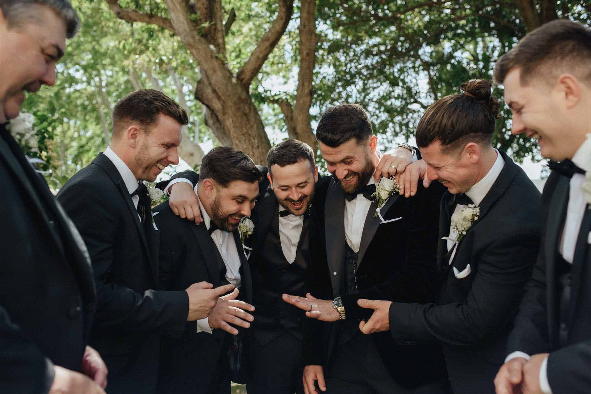 Groom and seven groomsmen in black suits with cream buttonholes gathered in a circle under trees in a garden.