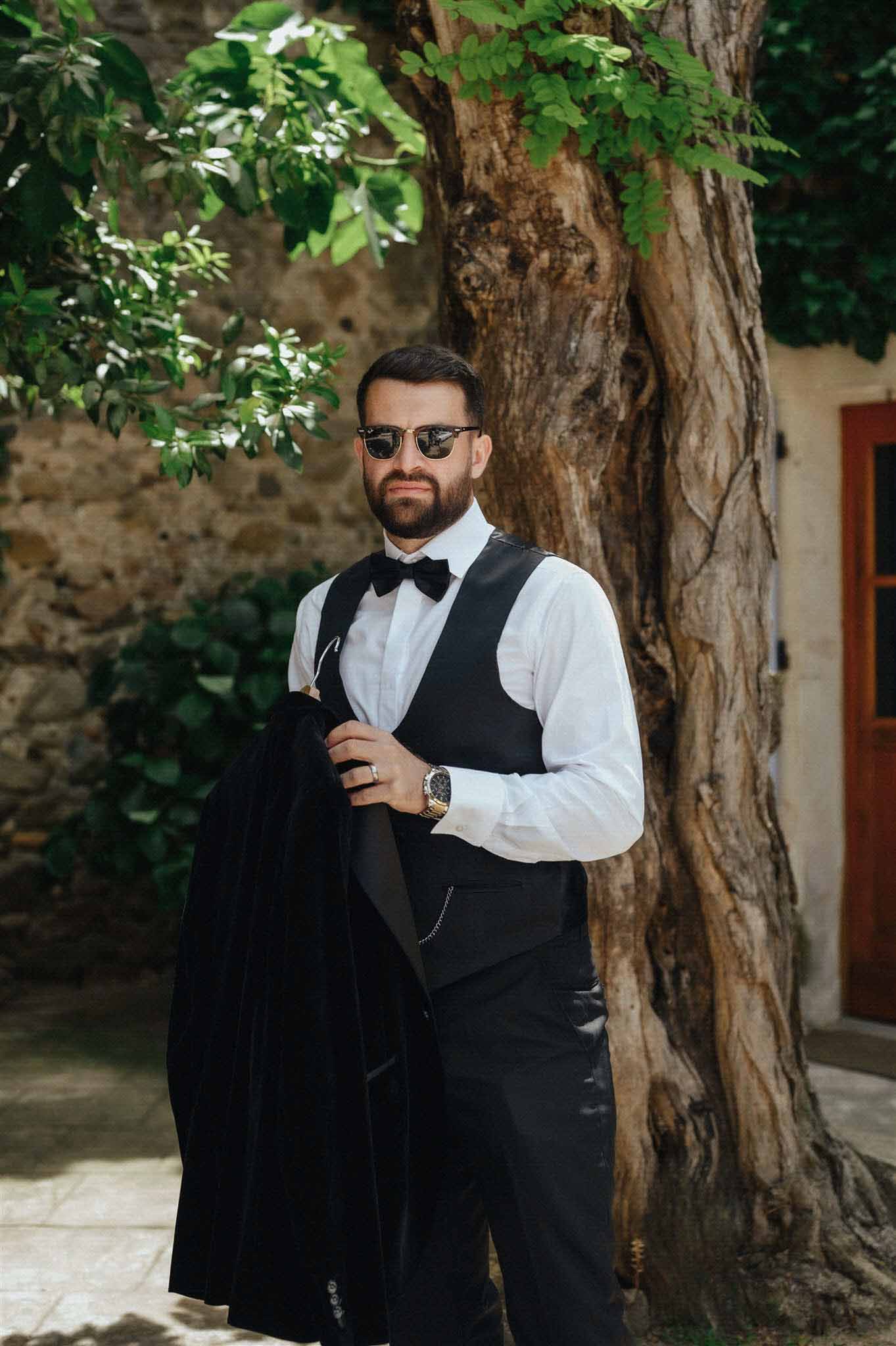 Groom in tuxedo holding jacket and wearing black sunglasses at Château du Puits-es-Pratx