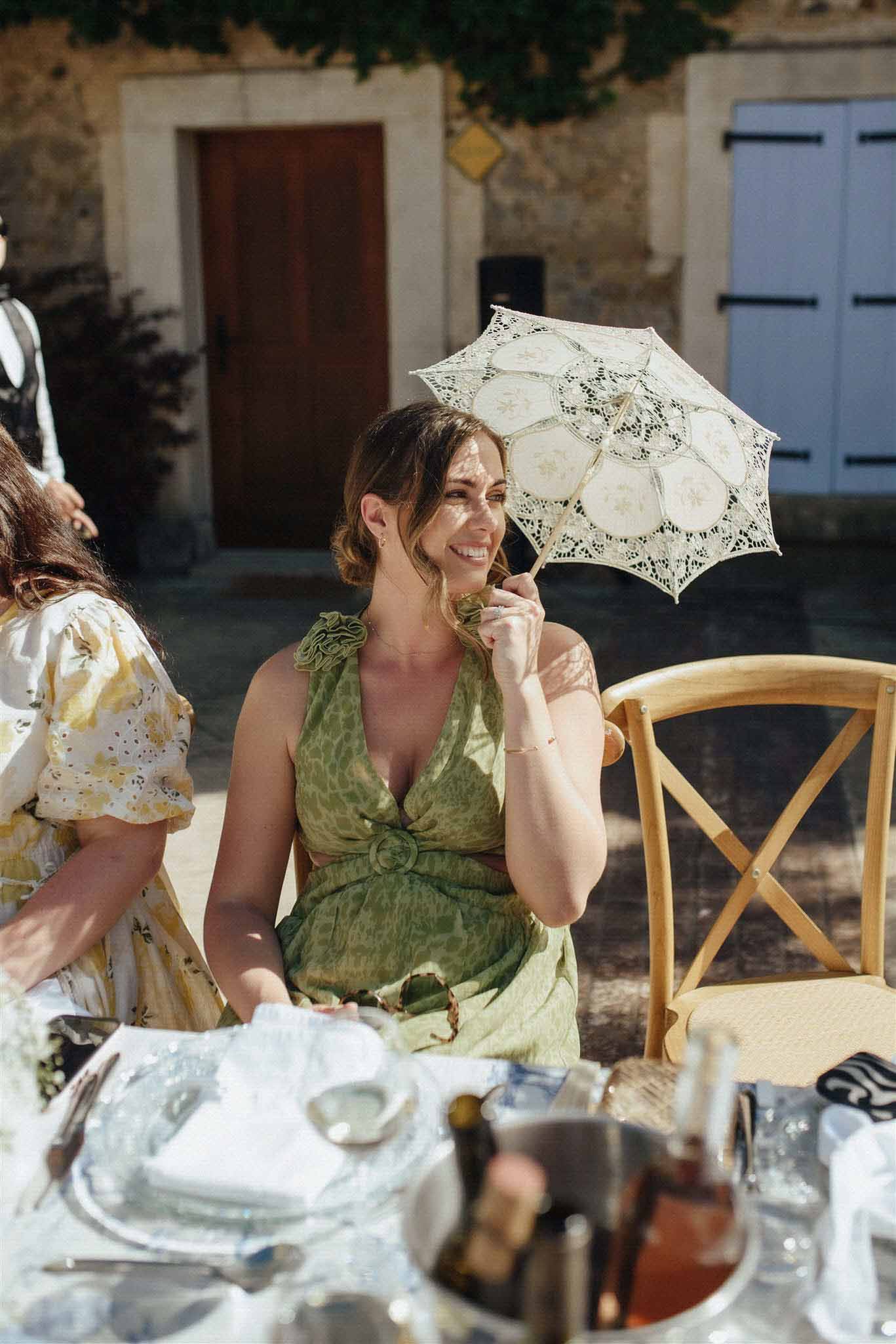 Guest in sage green dress holding cream parasol seated at outdoor reception table