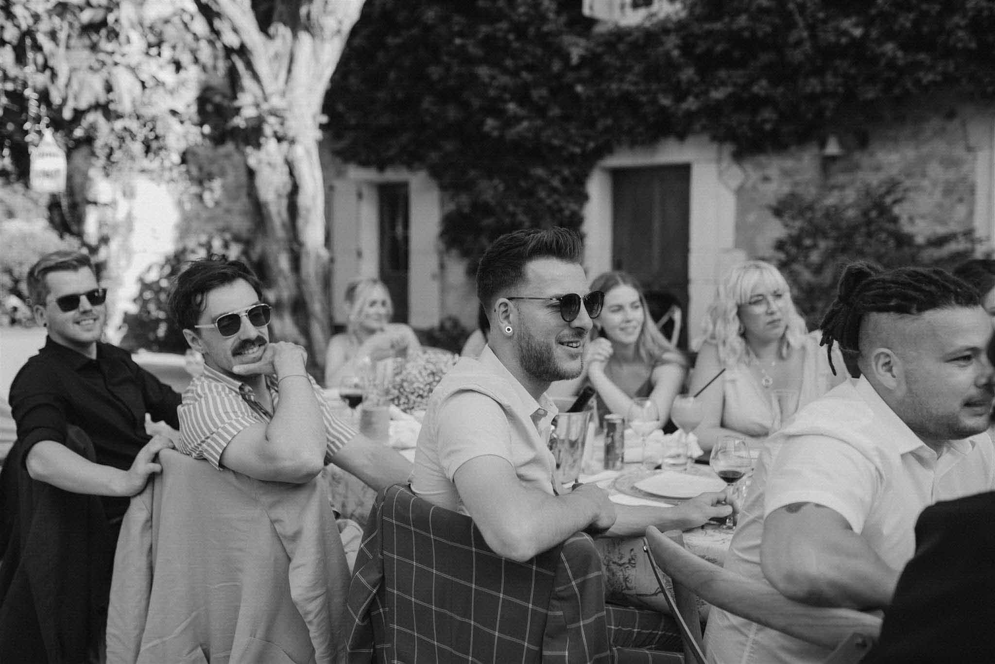 Black-and-white candid of reception guests seated at outdoor table with man in tartan kilt smiling
