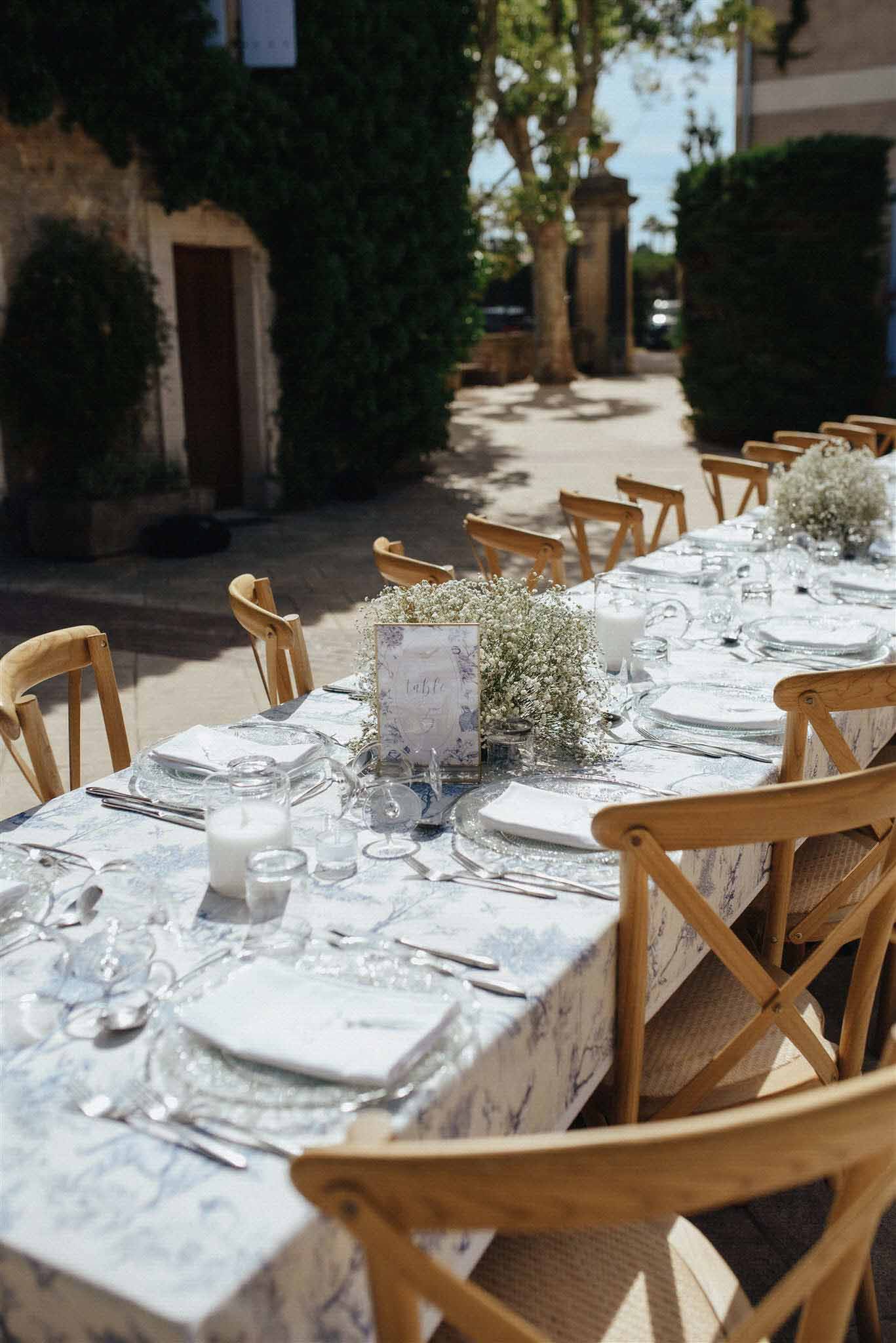 Long banquet table with pale blue linen, white plates, wooden cross-back chairs and baby's breath centerpieces in a stone courtyard.
