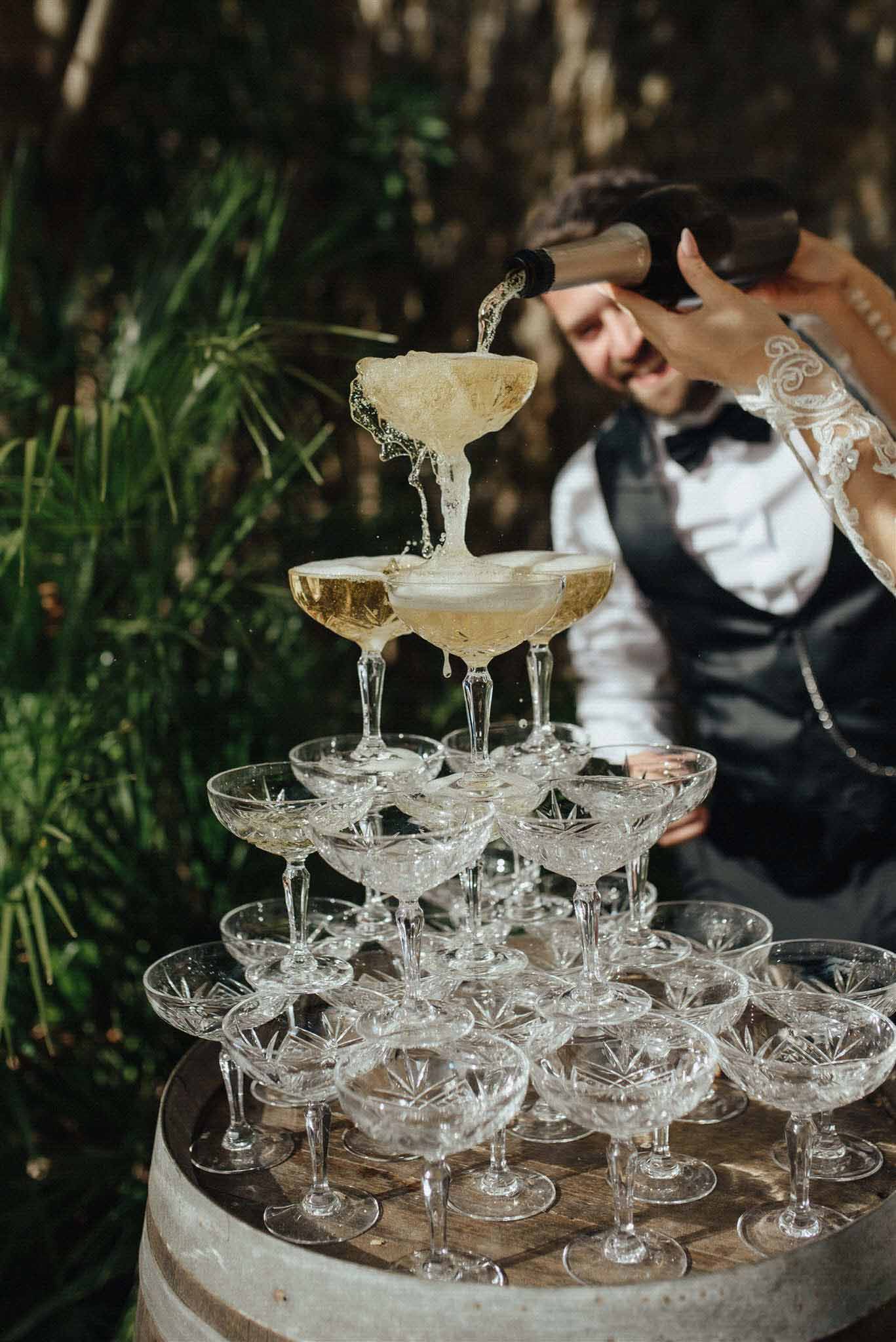 Groom pouring champagne into tiered crystal coupe tower outdoors, bride in white lace dress softly out of focus behind