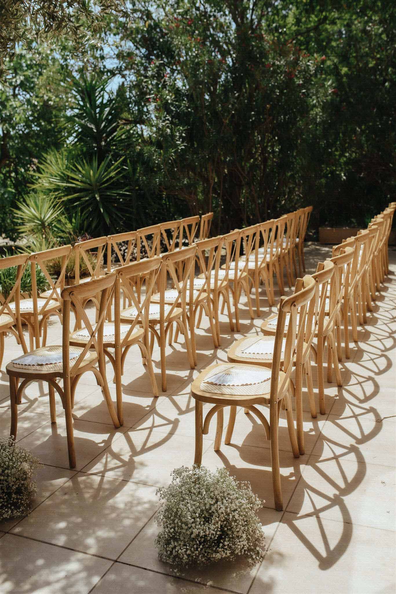 Outdoor ceremony setup with rows of wooden cross-back chairs on a garden terrace flanked by trees and greenery