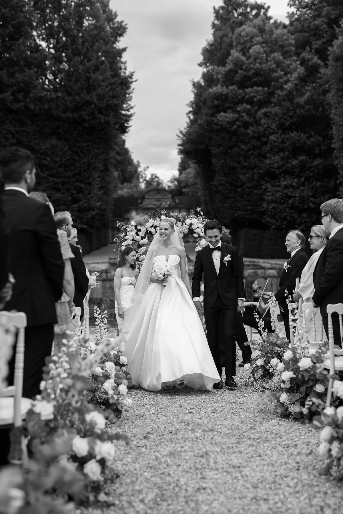 Black-and-white recessional with bride and groom walking down hedge-lined aisle