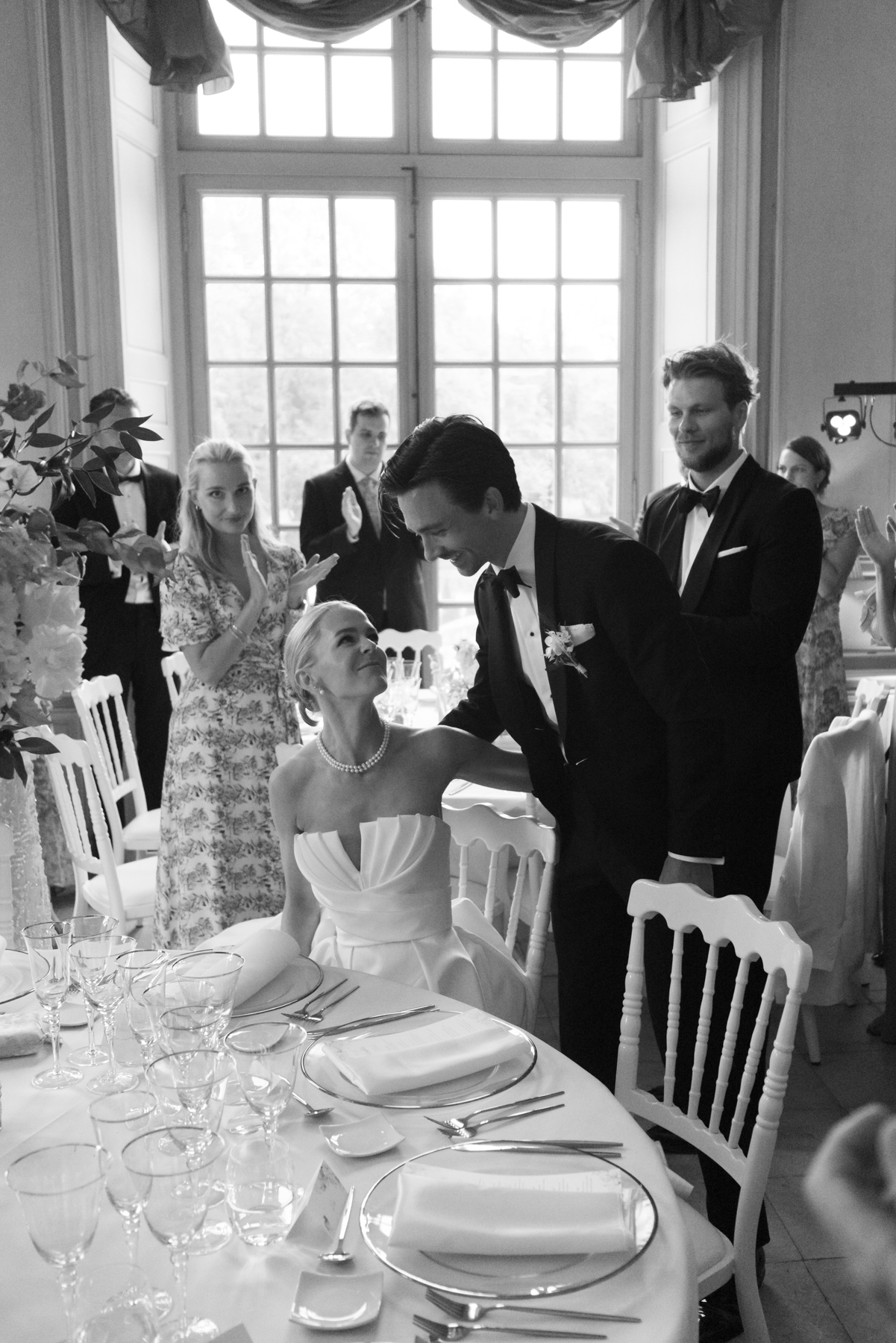 Black-and-white reception scene with groom in tuxedo standing over seated bride at formally set dinner table