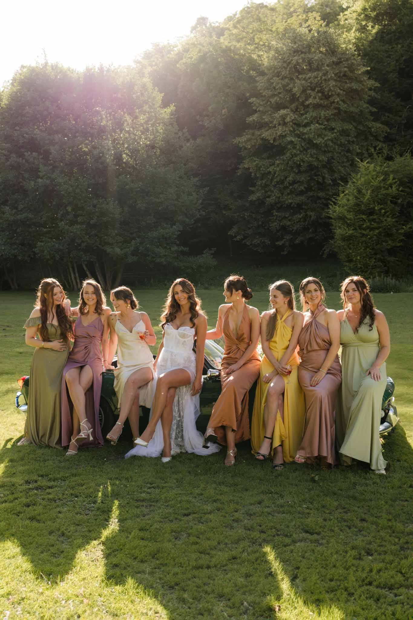 Bride with eight bridesmaids in coordinated earth tone dresses on golden hour lawn