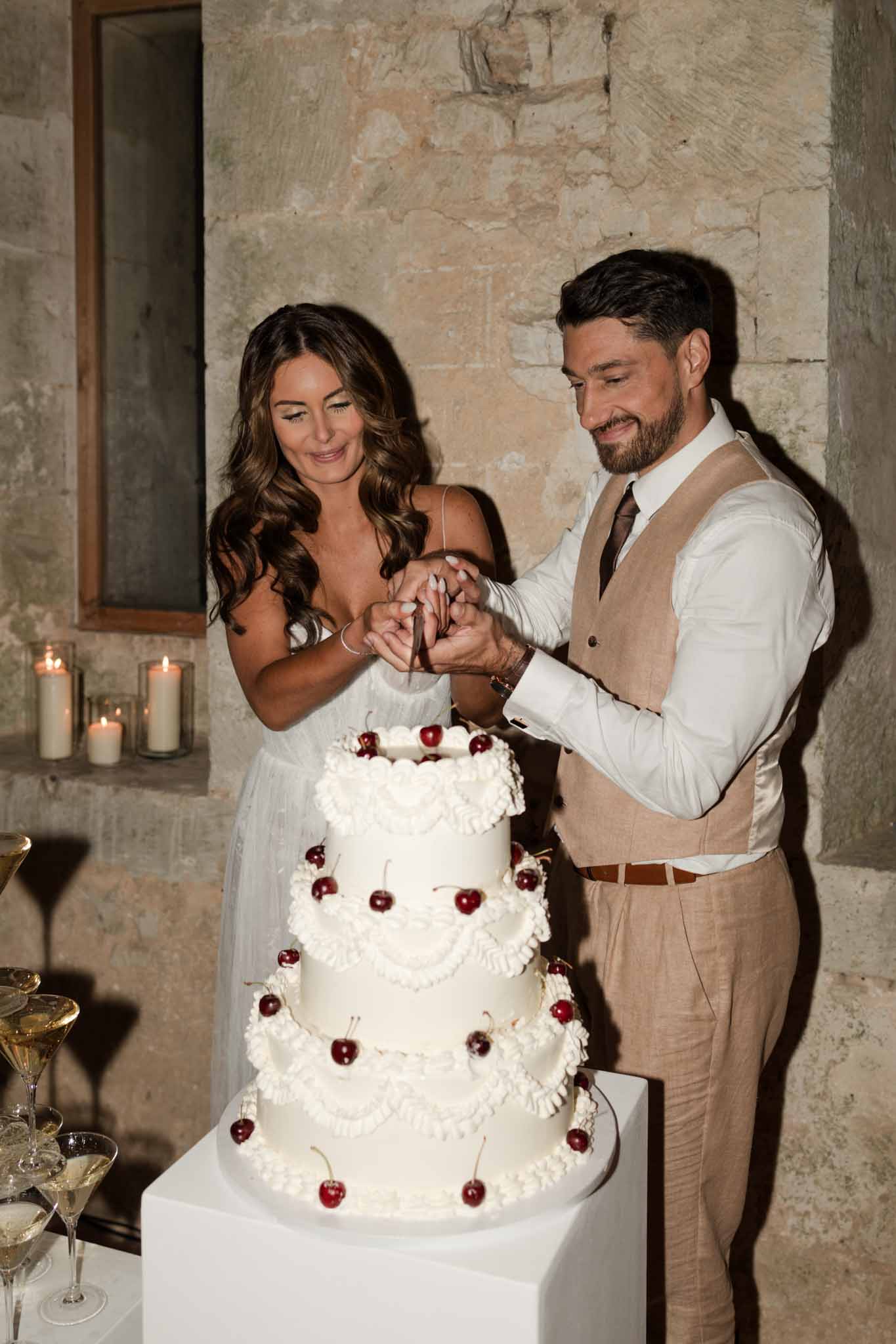 Bride and groom cutting a three-tiered ivory cake decorated with burgundy cherries against a rustic stone wall