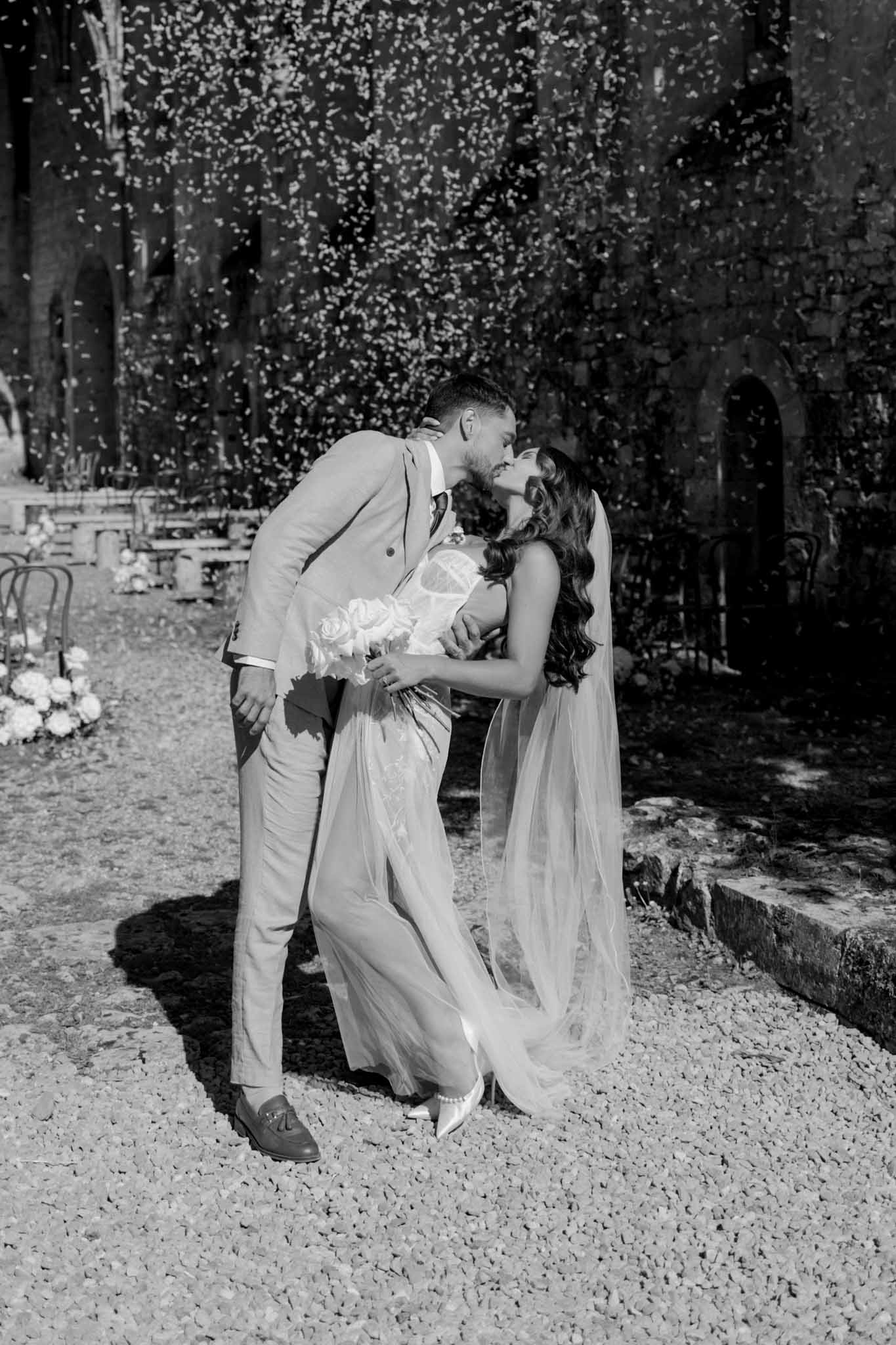 Black-and-white portrait of groom kissing bride on gravel pathway with ivy-covered walls in garden courtyard