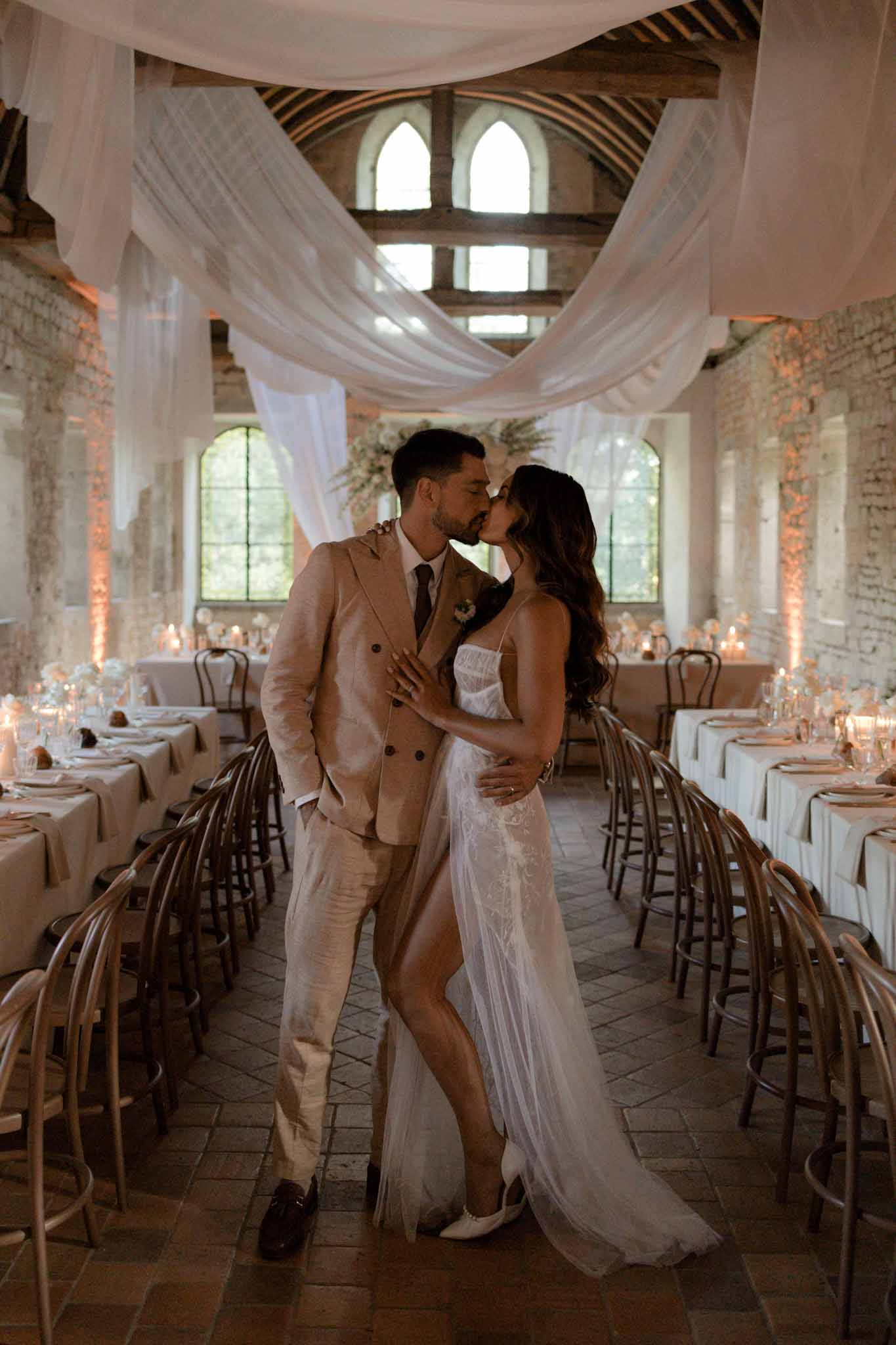 Bride in white lace dress and groom in tan suit kiss down center aisle of brick-walled venue with sheer ceiling draping