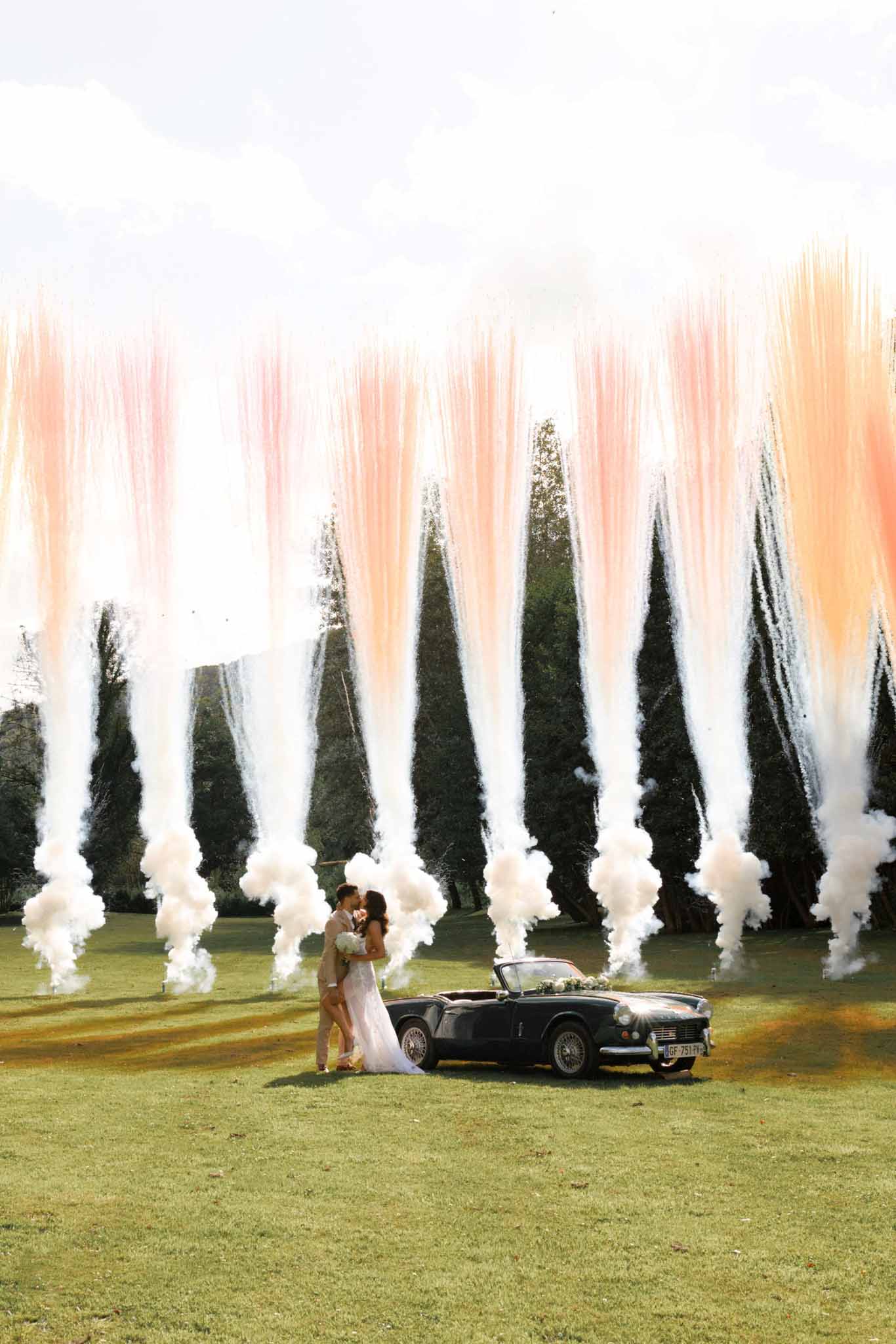 Bride and groom kissing beside vintage convertible with white smoke and cypress trees