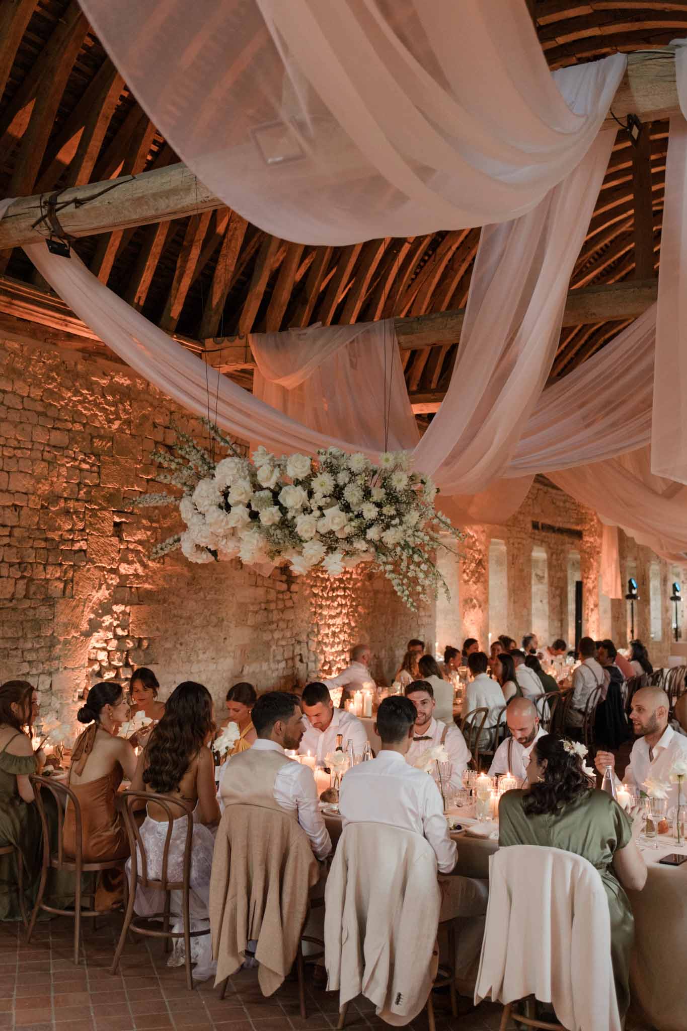 Reception in rustic brick venue with exposed beams, blush fabric draping, white floral installation above long tables