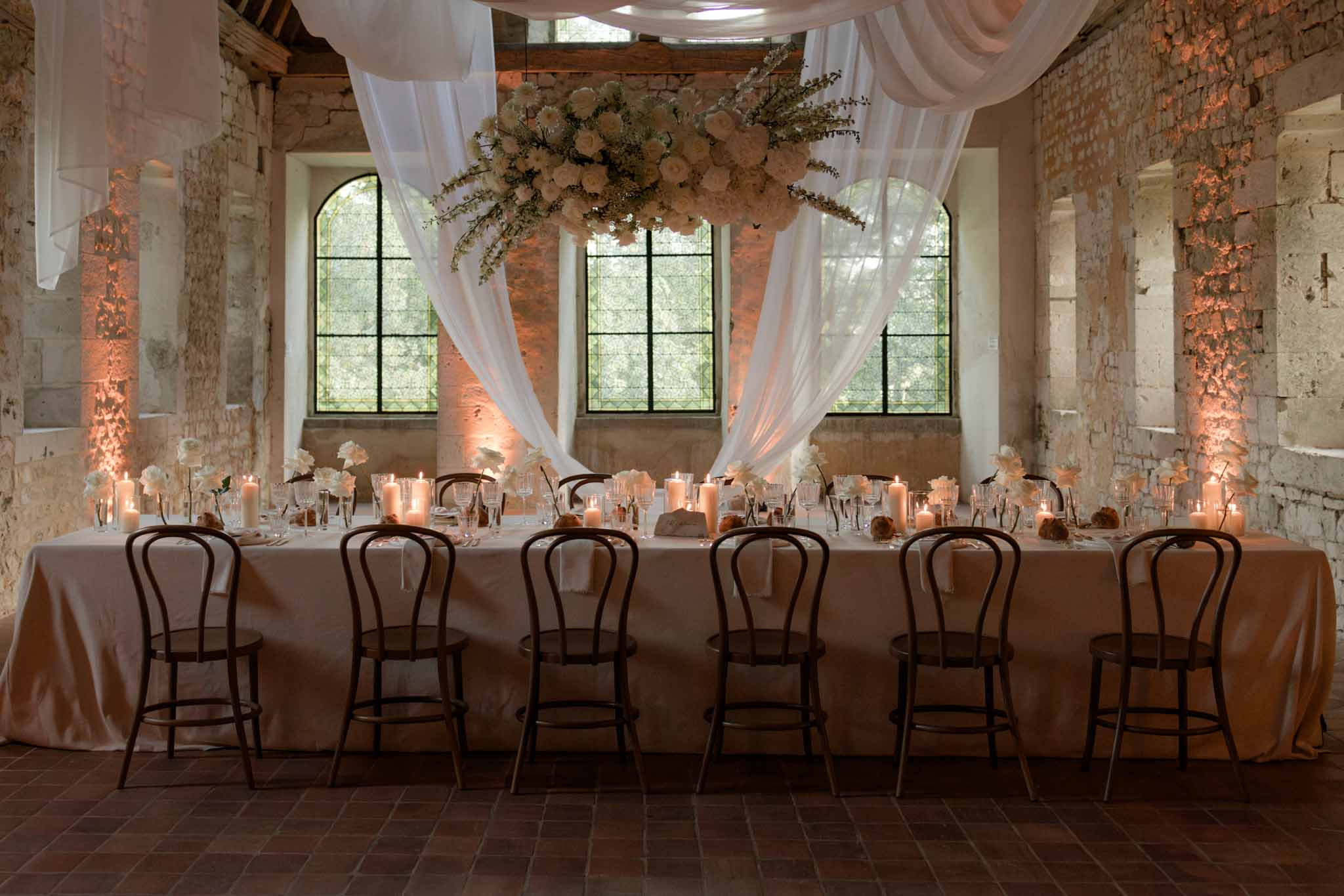 Long reception table in industrial brick venue with suspended cream floral arrangement, candles and sheer draping