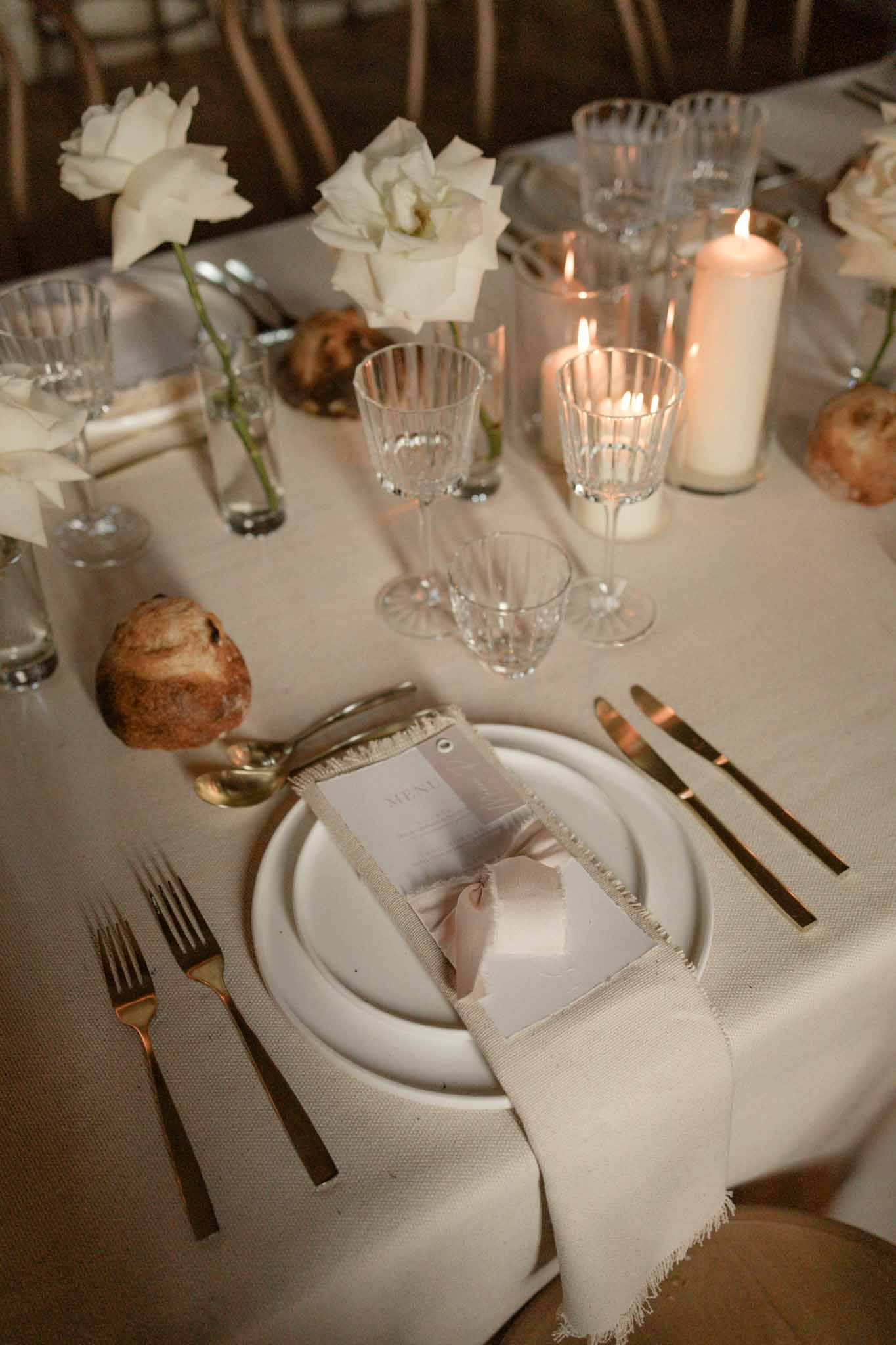 Reception place setting with cream linen, gold flatware, white gardenias, pillar candles, and menu card