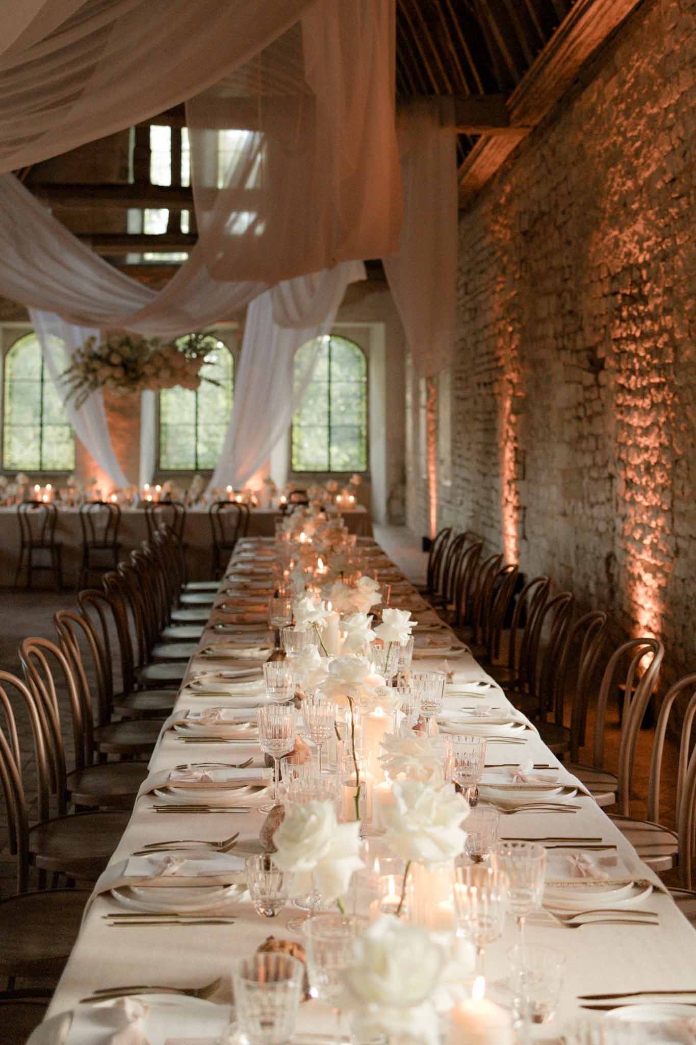 Long banquet table with cream linen, white florals and candles in brick-walled venue with sheer ceiling draping