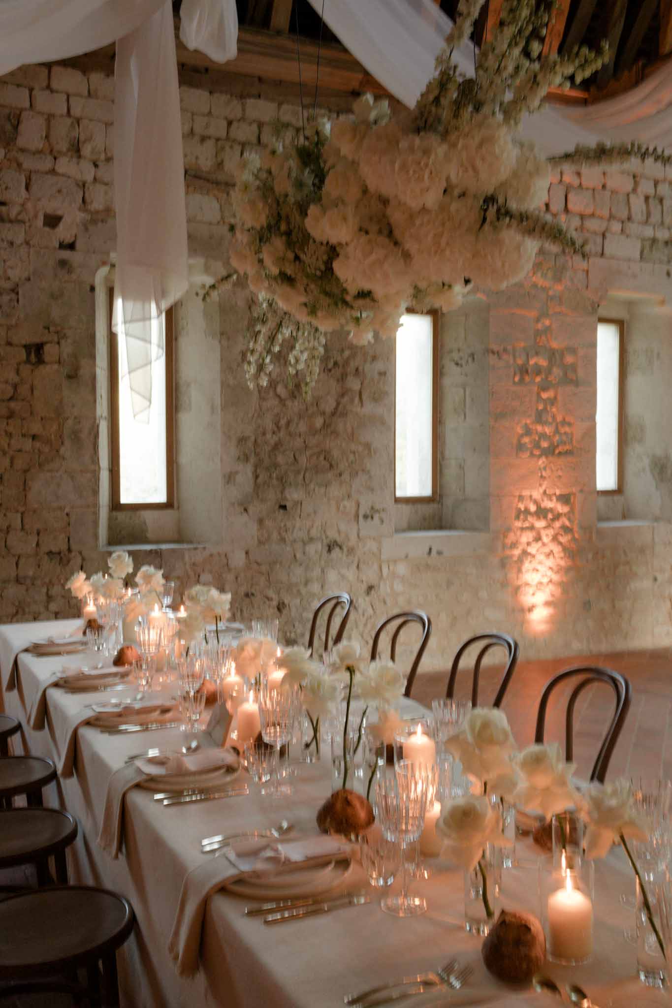 Long rustic stone-walled reception venue with arched windows, cream florals, and dried greenery ceiling installation