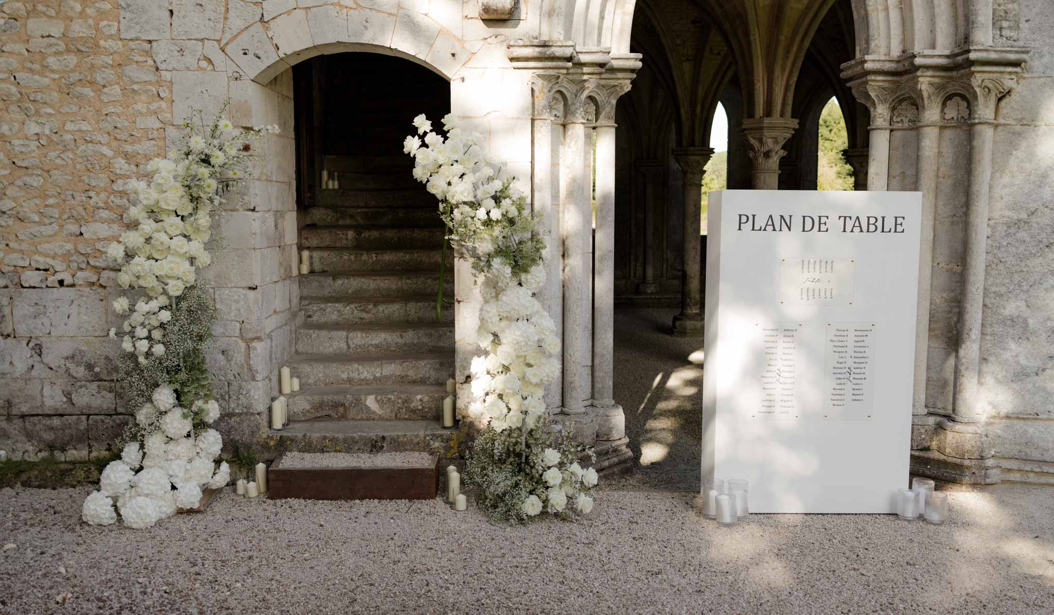 Seating chart in stone cloister corridor flanked by white rose arrangements on wooden stands with pillar candles
