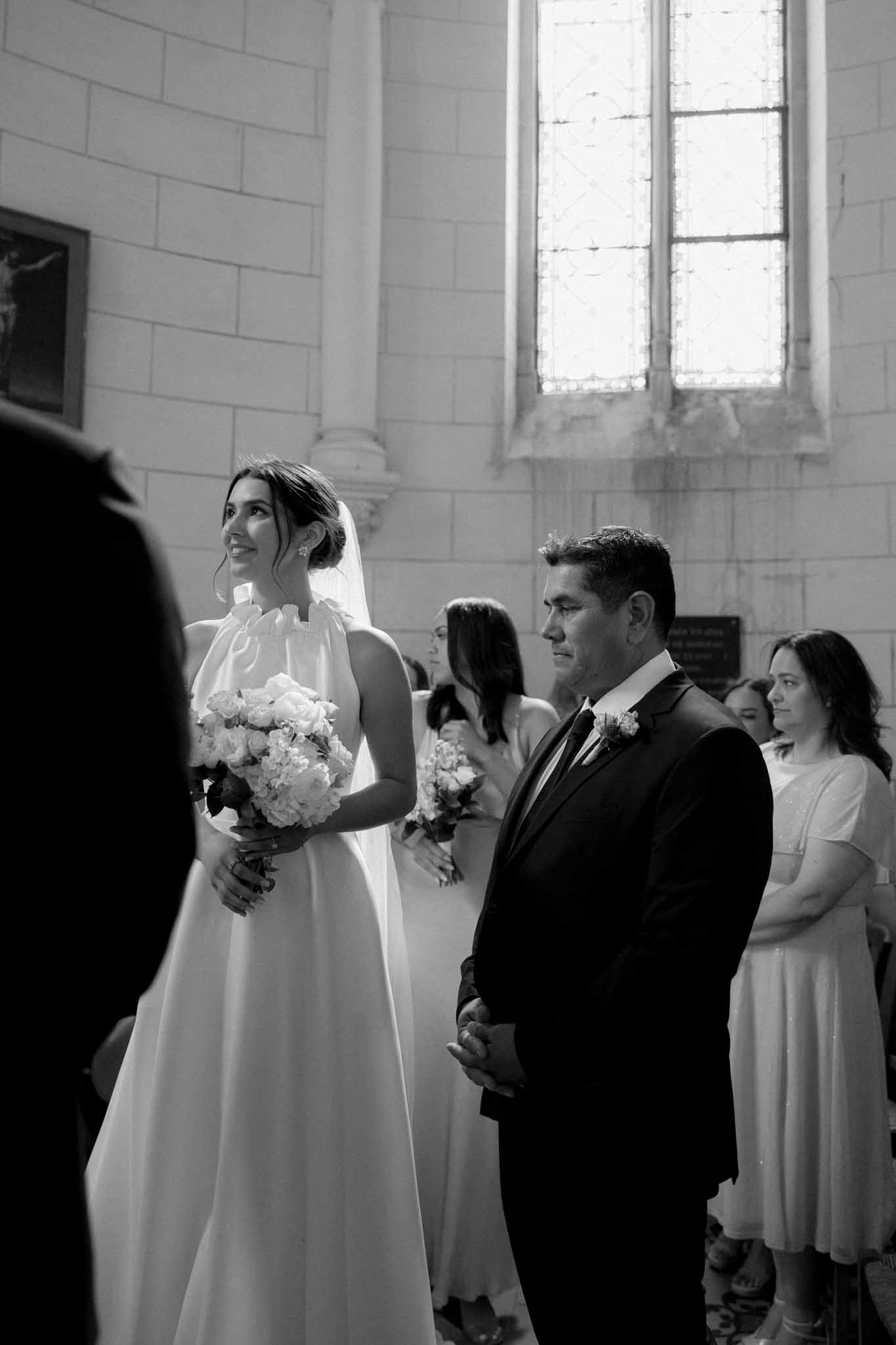 Bride with white bouquet and groom in dark suit during ceremony in stone chapel with arched windows, black-and-white