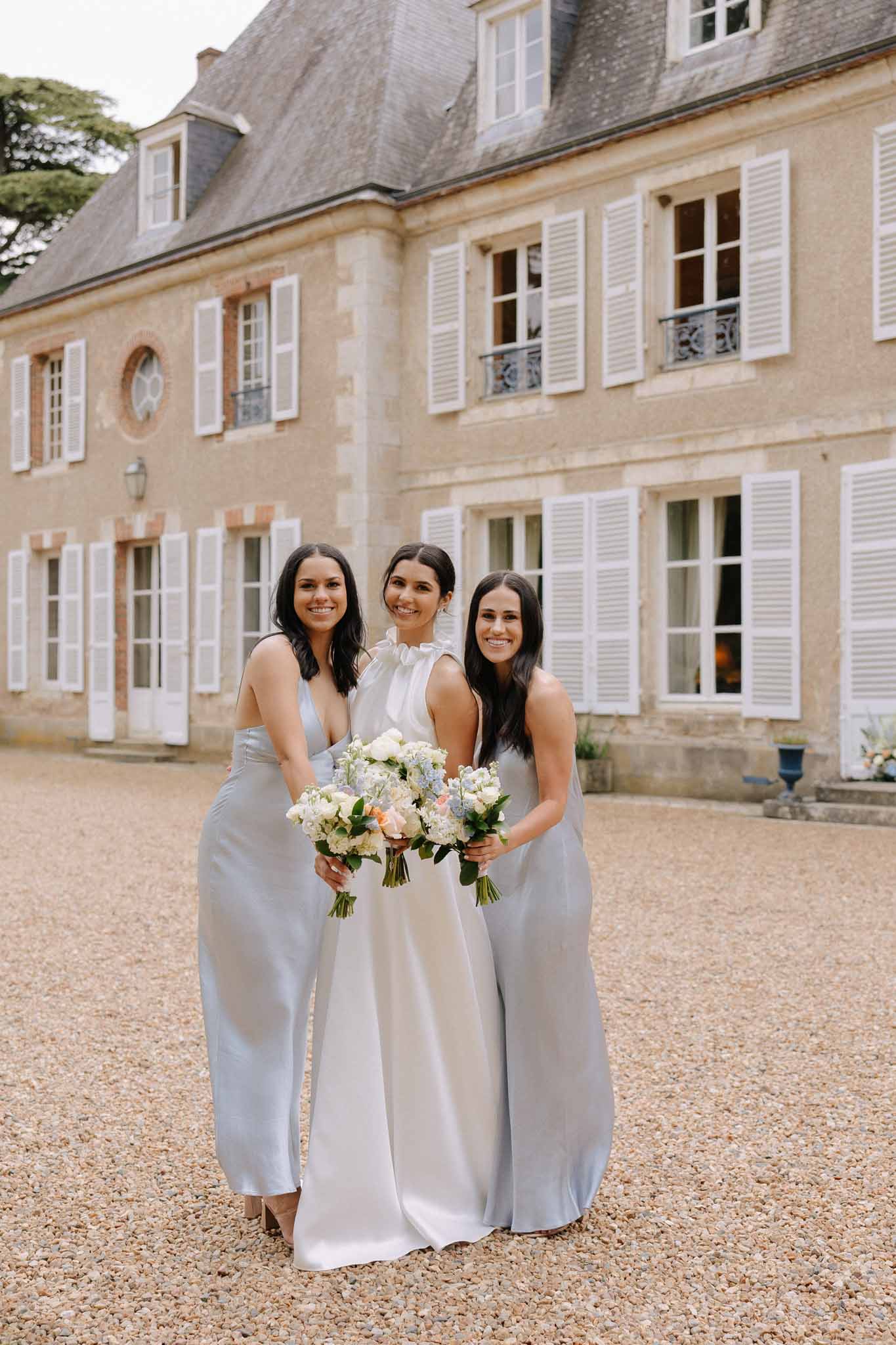 Bride in white gown with two bridesmaids in grey dresses standing in courtyard of French stone manor house