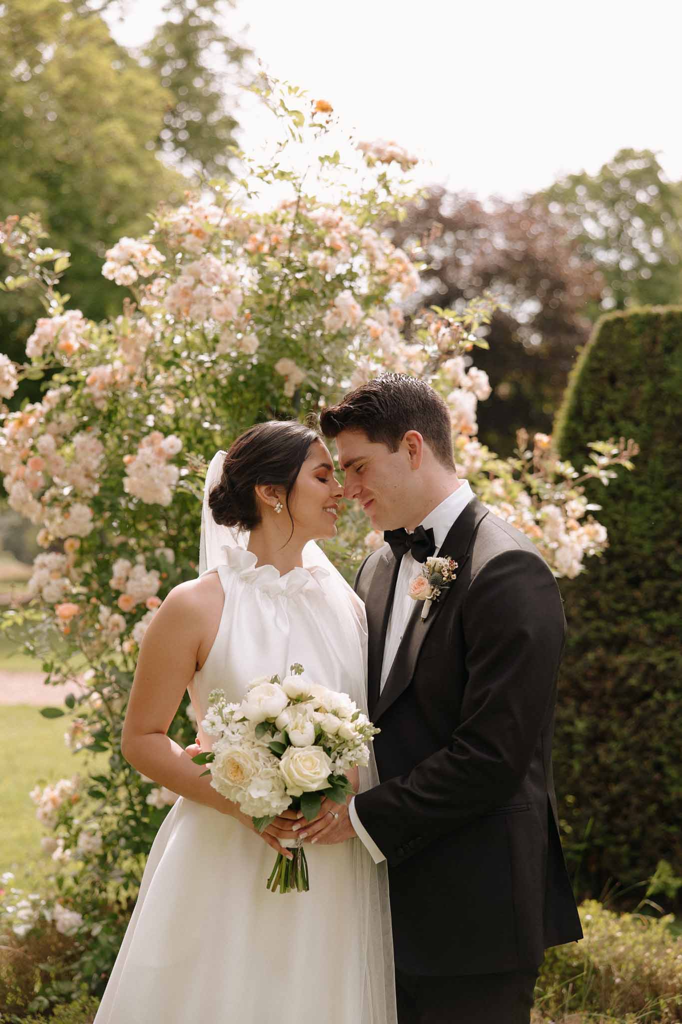 Bride in white sleeveless gown with veil and groom in black tuxedo in close portrait, climbing pink rose vine as garden backdrop
