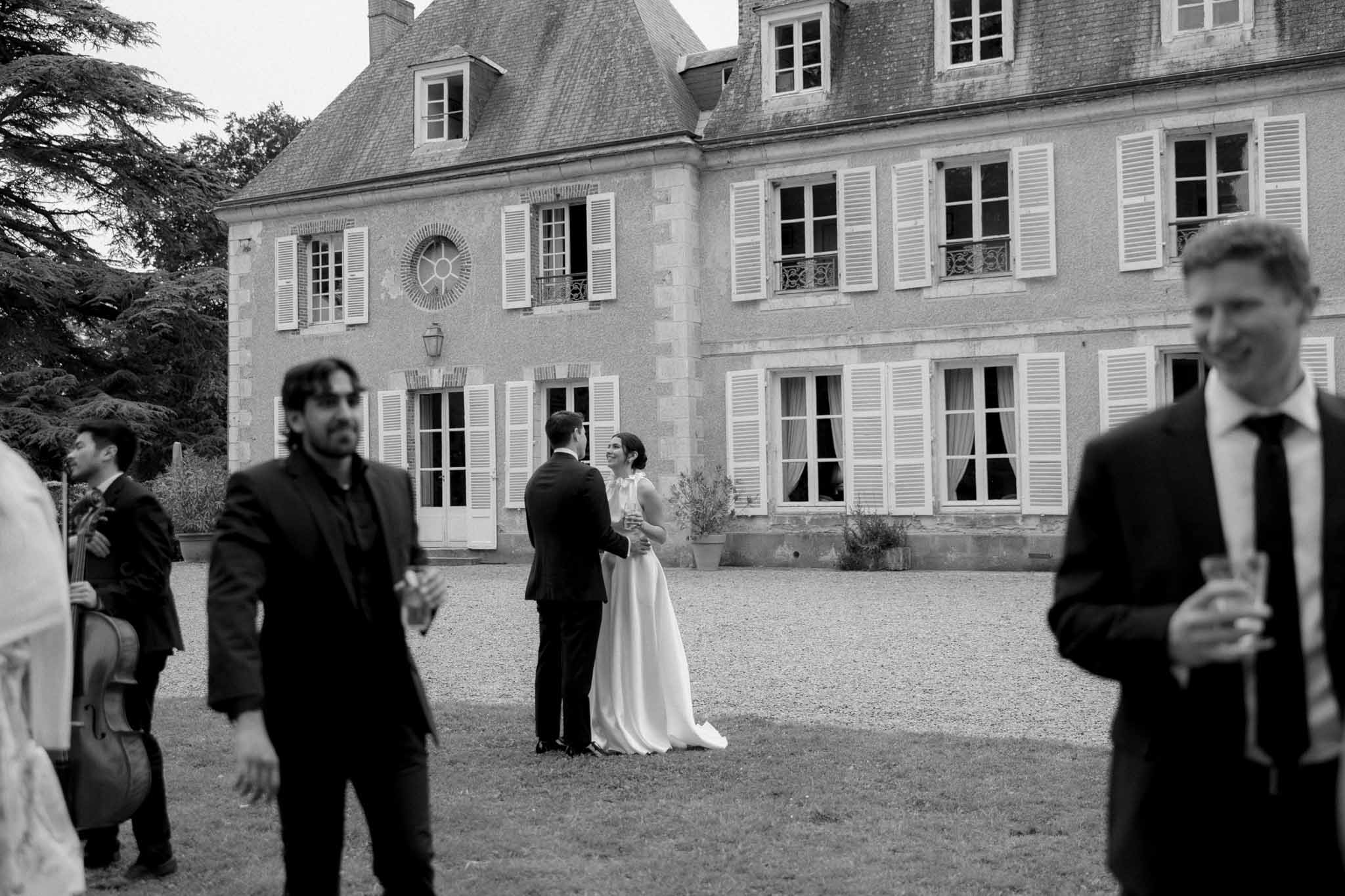 Black-and-white photo of bride and groom conversing in gravel courtyard of French manor house during cocktail hour