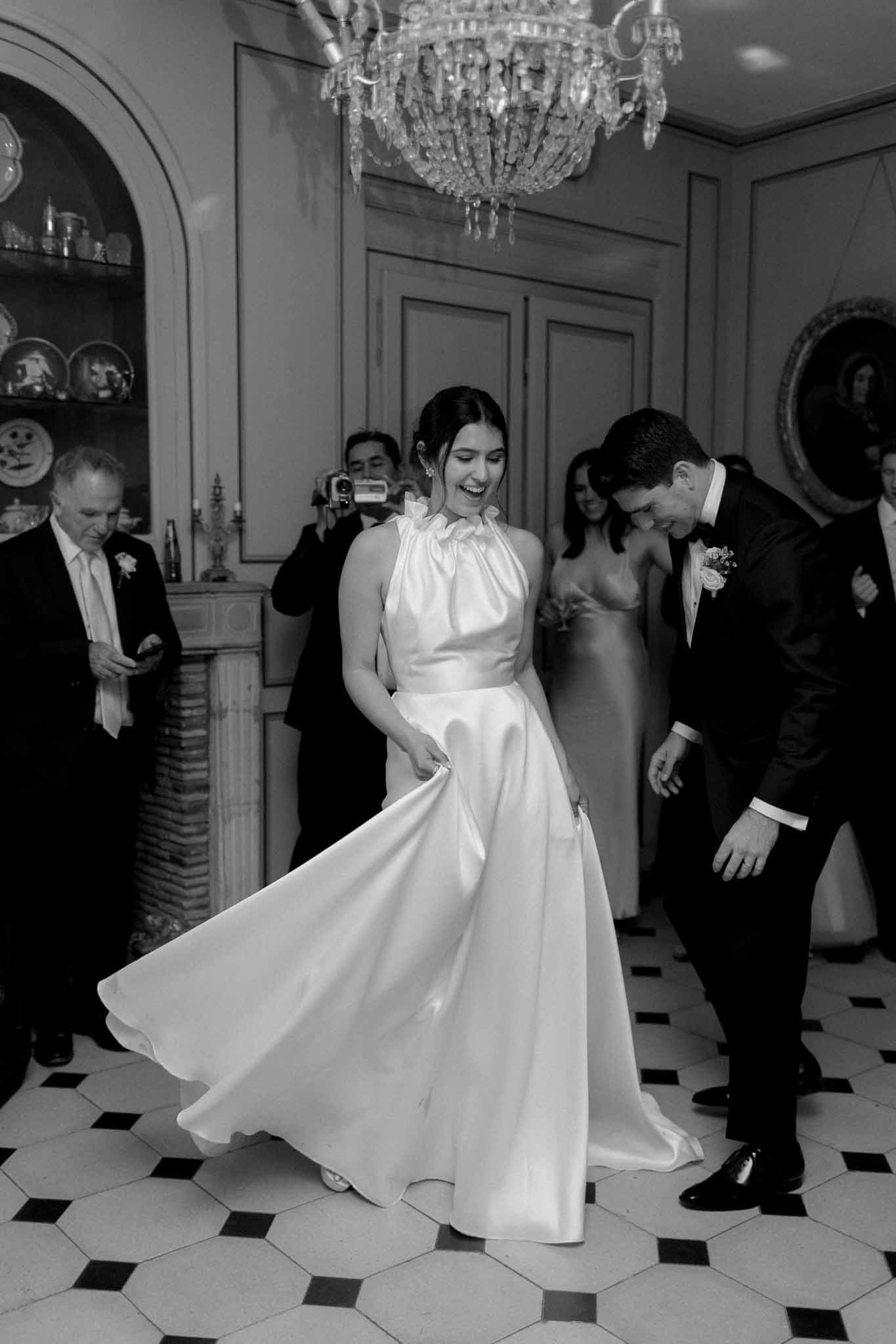 Bride in white halter-neck gown and groom in black tuxedo in ornate ballroom, black and white, guests watching