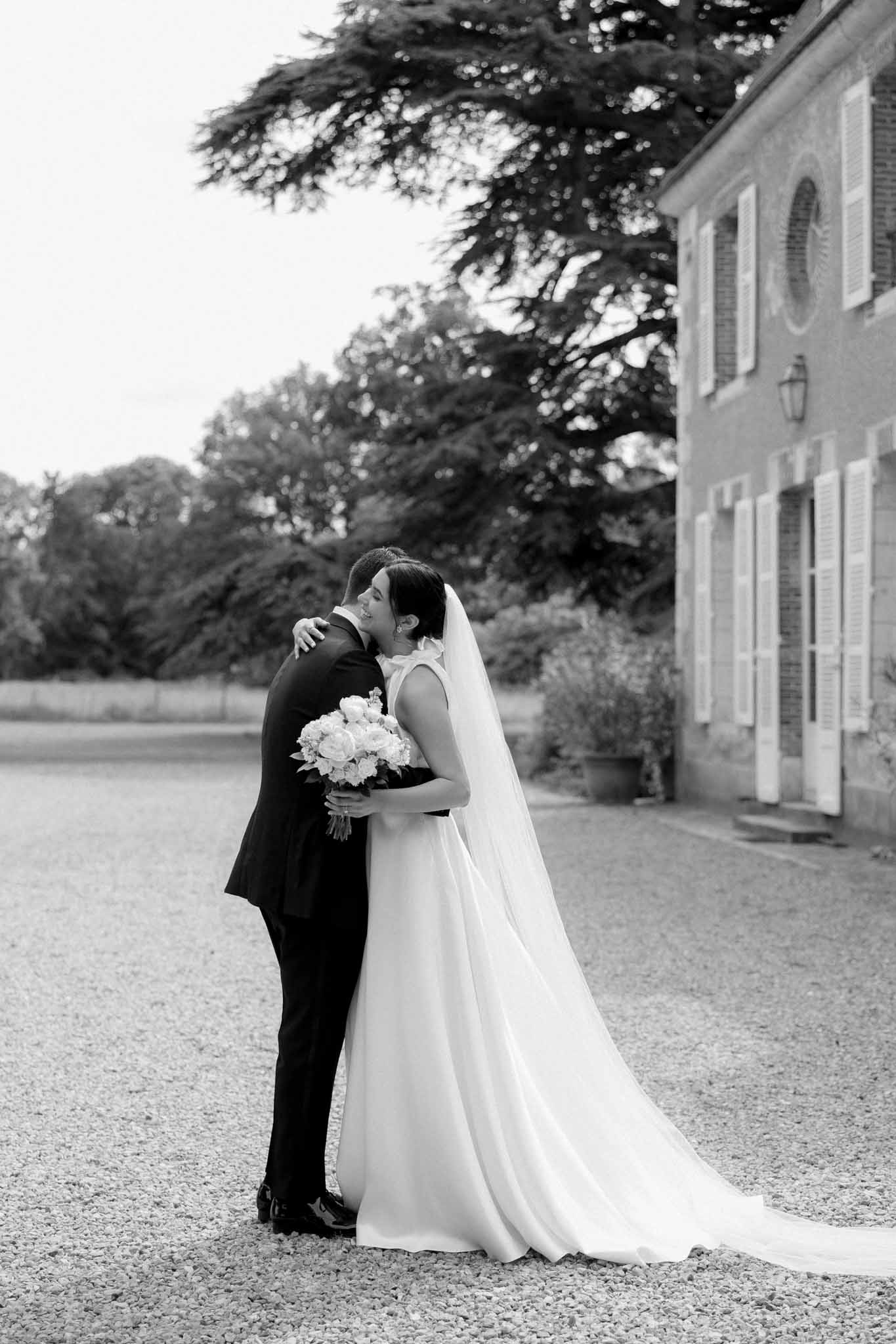 Black and white portrait of groom embracing bride from behind on gravel drive, brick manor house and trees in background
