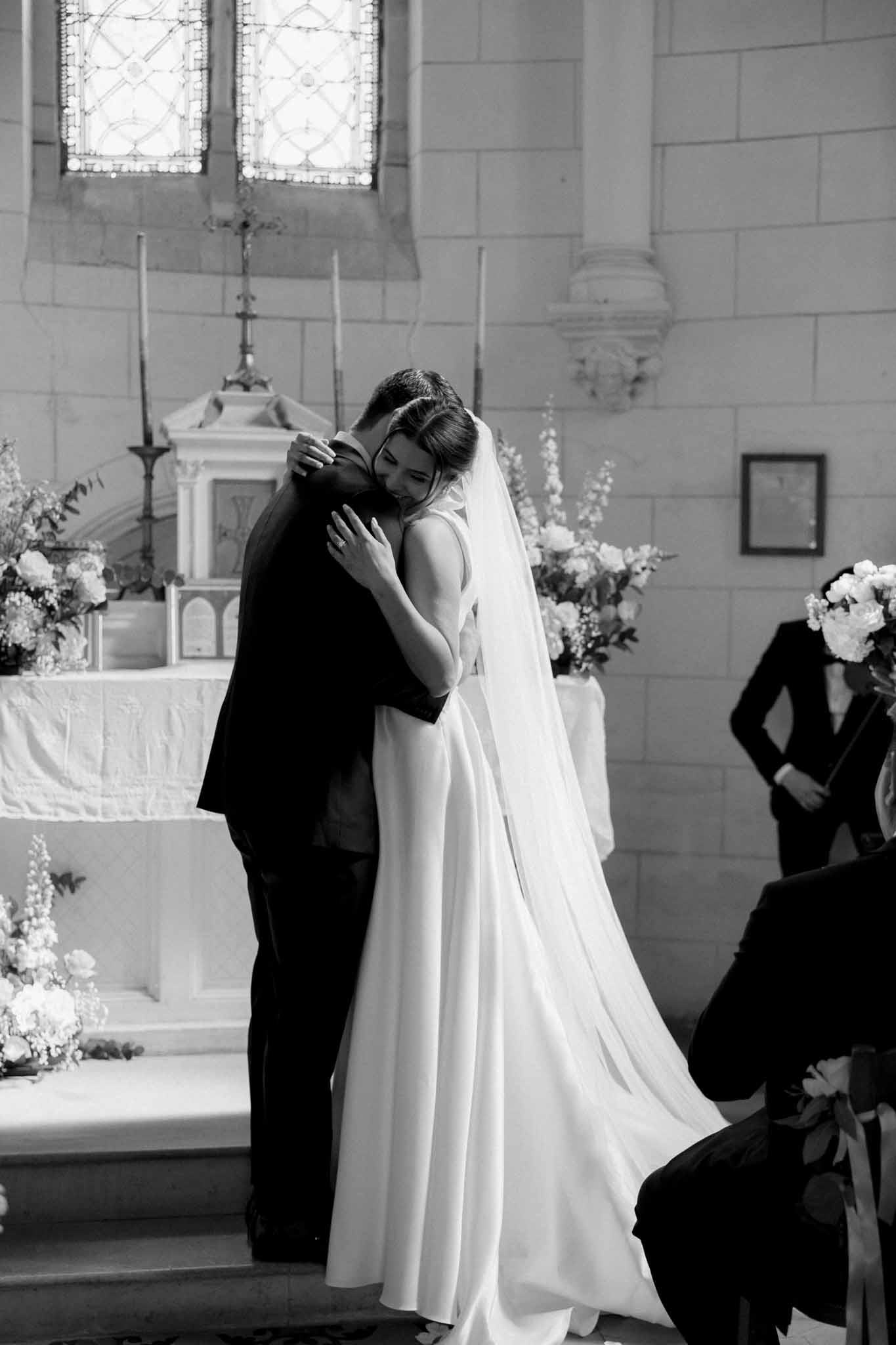 Couple embracing at the chapel altar with white floral arrangements and tall white candles on either side.