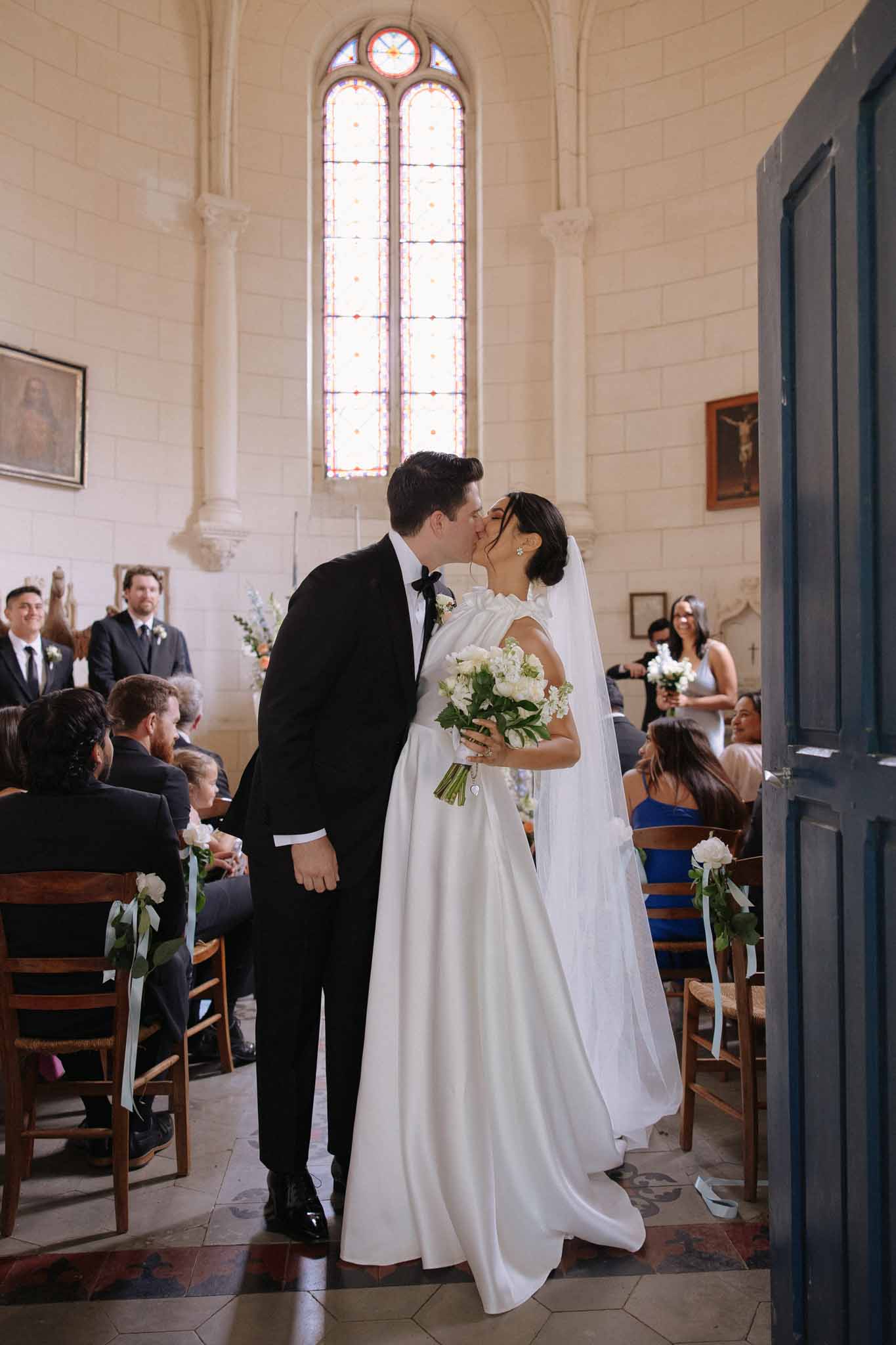 Bride and groom sharing their first kiss during a recessional in a stone chapel with stained-glass windows and white pew arrangements.