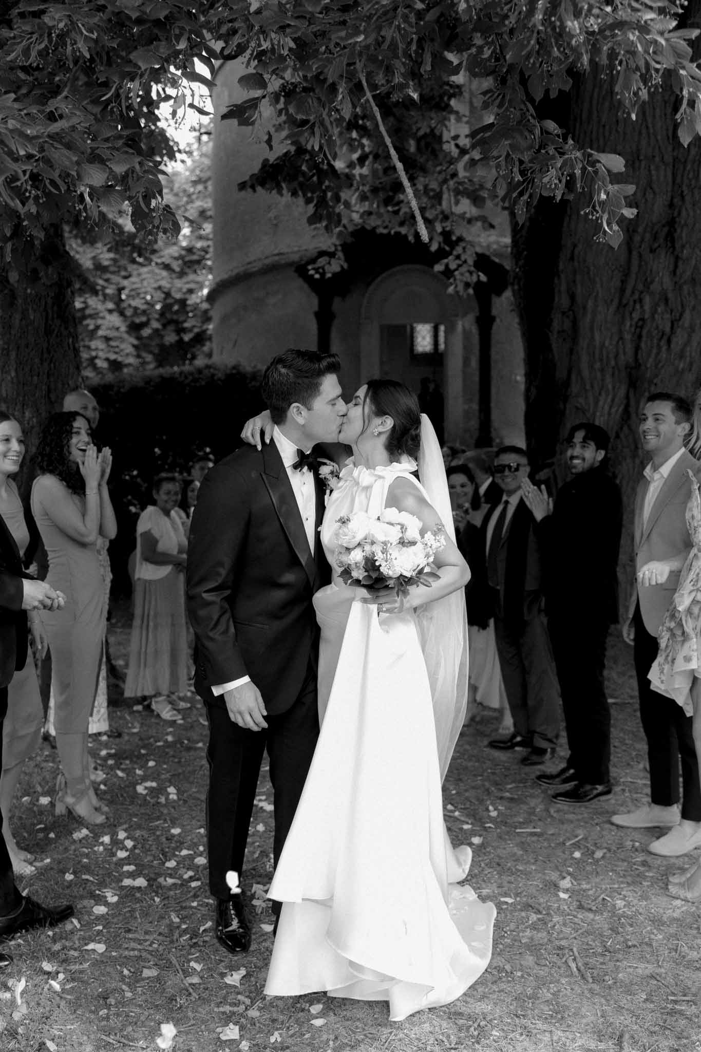 Black and white photo of bride and groom kissing in stone courtyard during recessional, guests applauding around them
