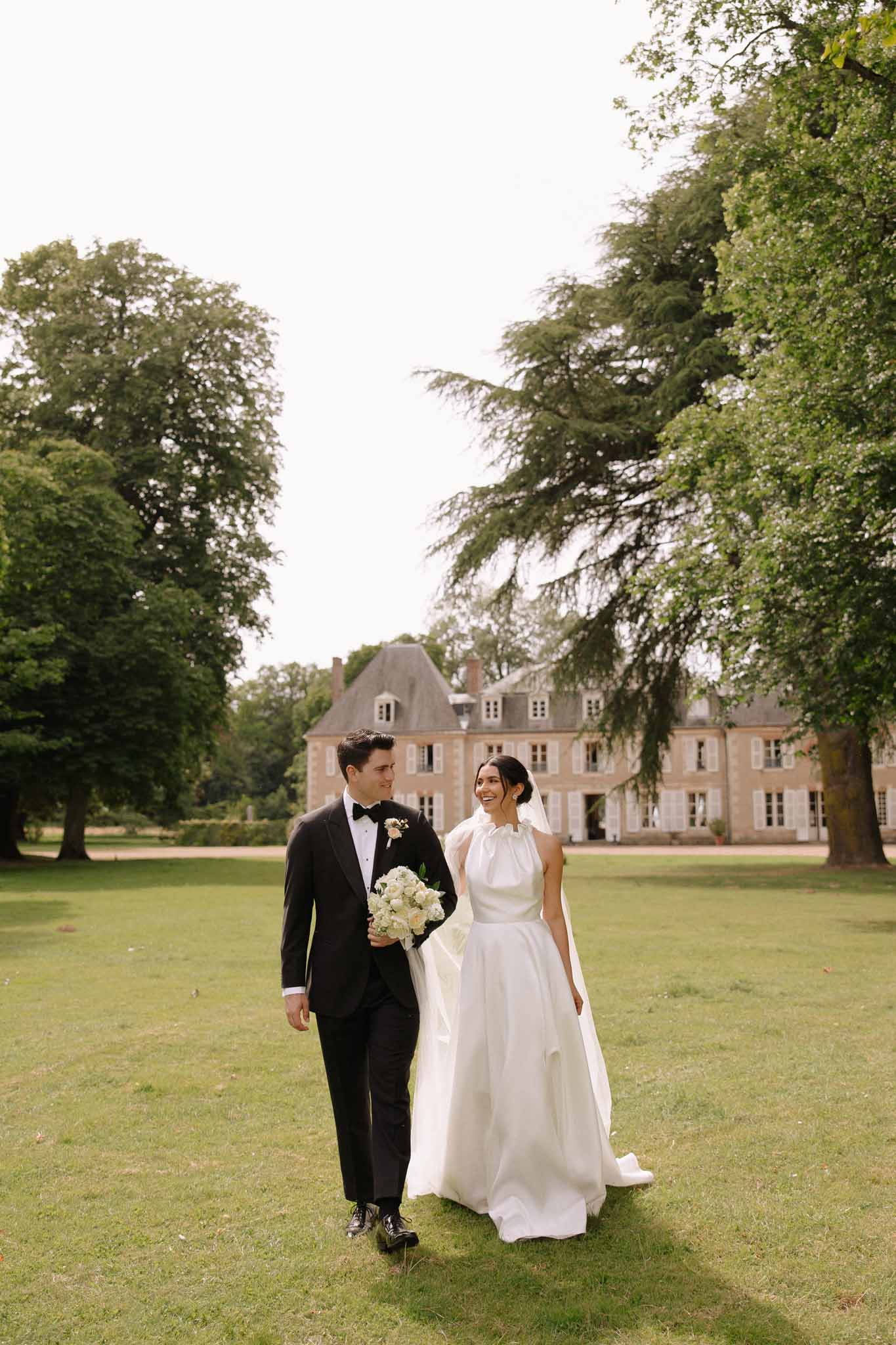 Bride in ivory gown and groom in tuxedo walking on manicured lawn in front of stone French château