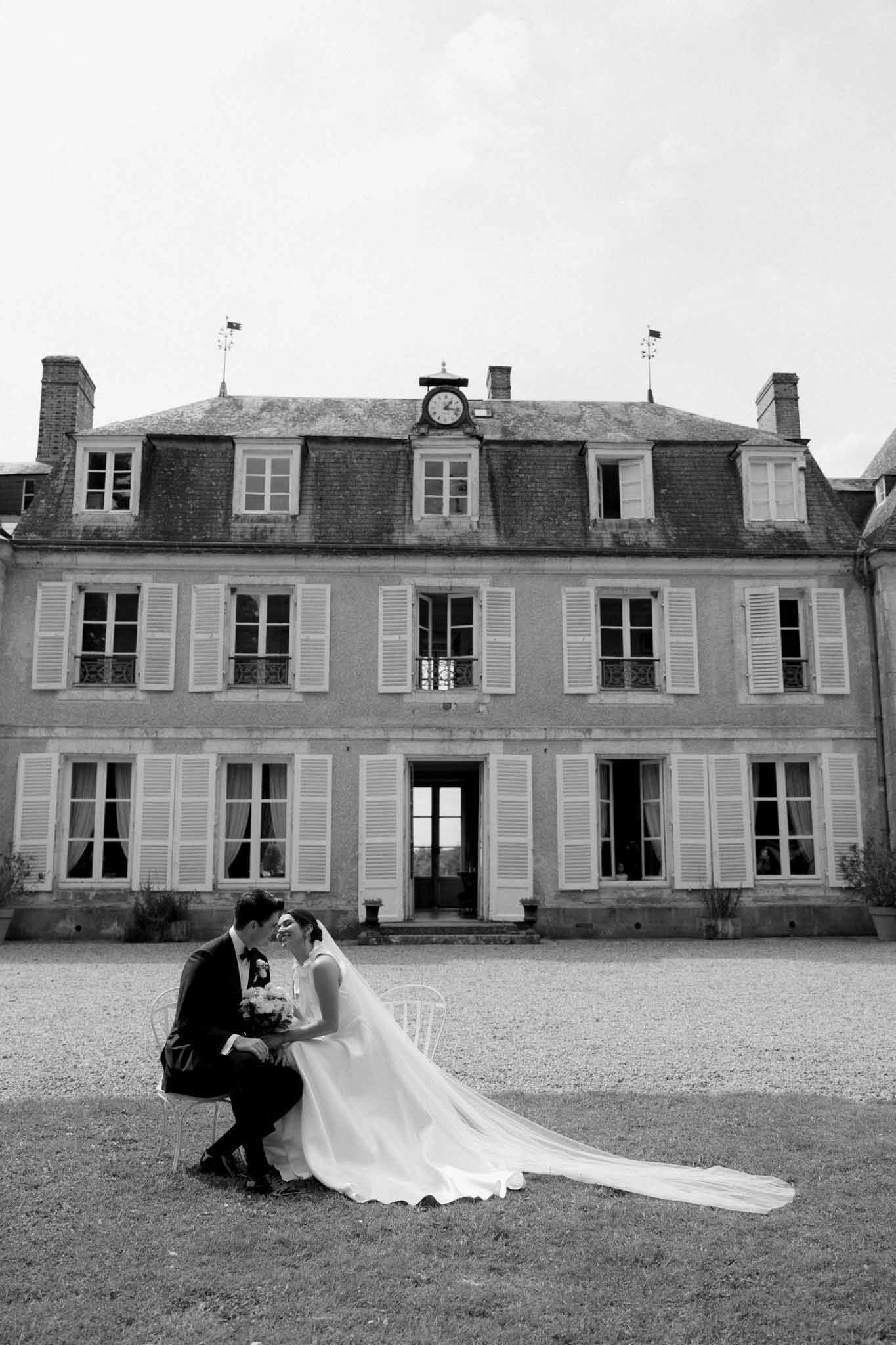 Black and white portrait of couple on Chiavari chair before three-story stone manor with clock tower, bride in white gown