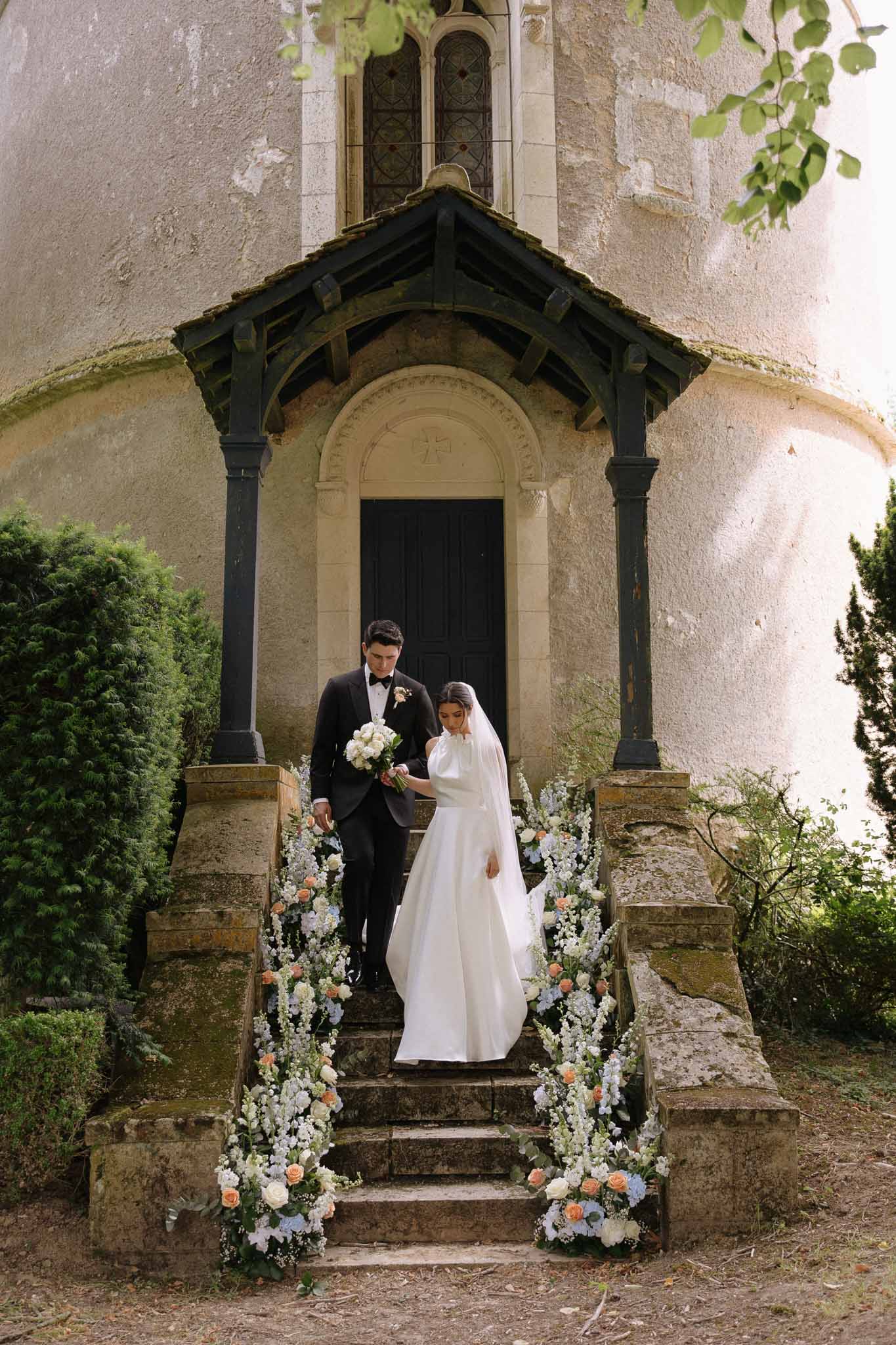 Bride and groom walking down chapel stairs flanked by floral arrangements at Château de Bouthonvilliers