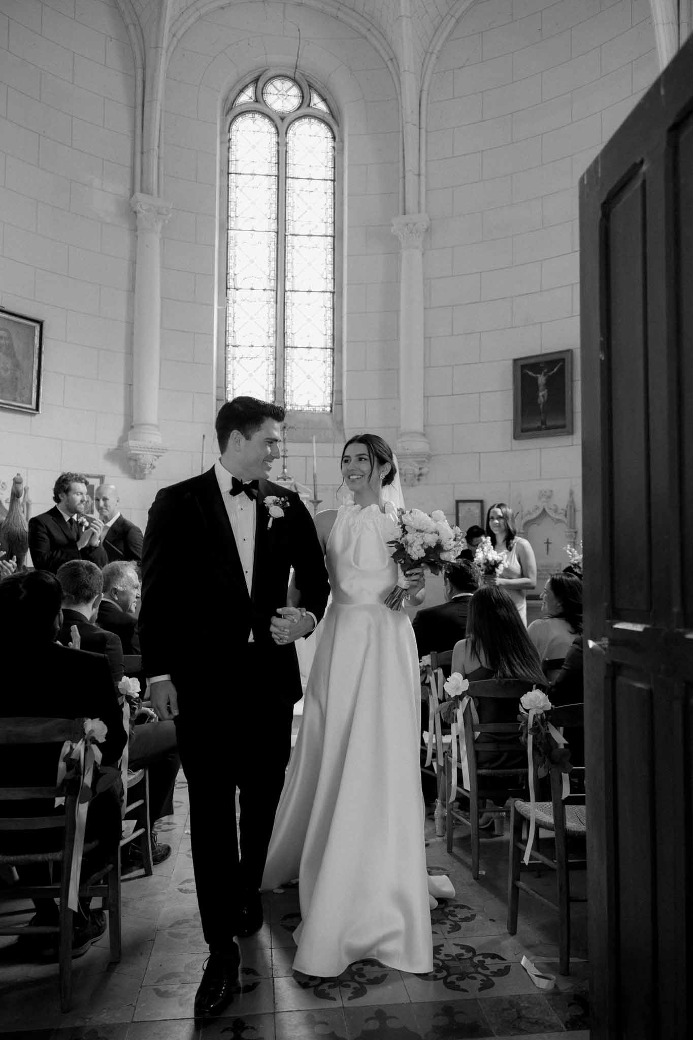 Black and white photo of bride and groom walking down chapel aisle past guests with stained glass above