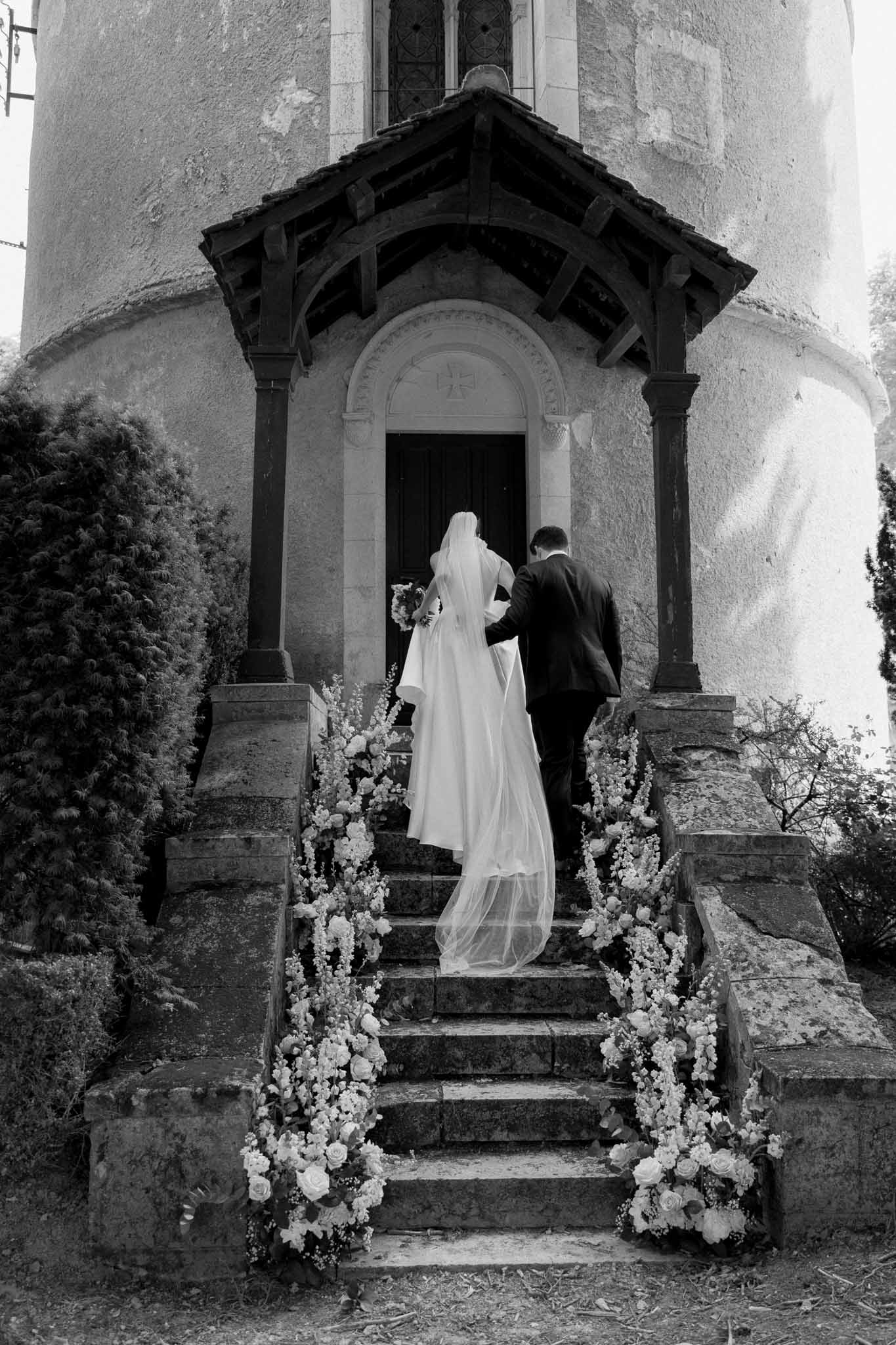 Black-and-white photo of bride and groom ascending stone chapel steps lined with white floral arrangements